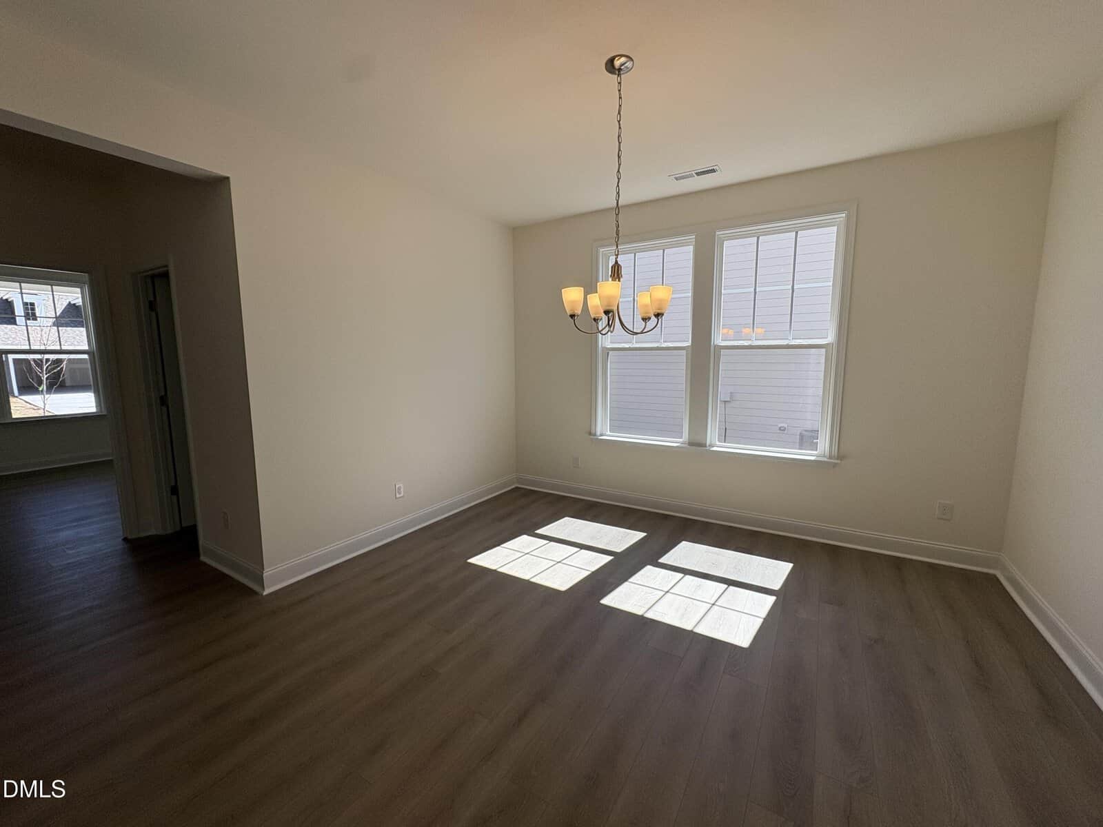A bright, empty room with light-colored walls, wood flooring, a chandelier, and two windows letting in sunlight. An open doorway leads to another room in the left background.
