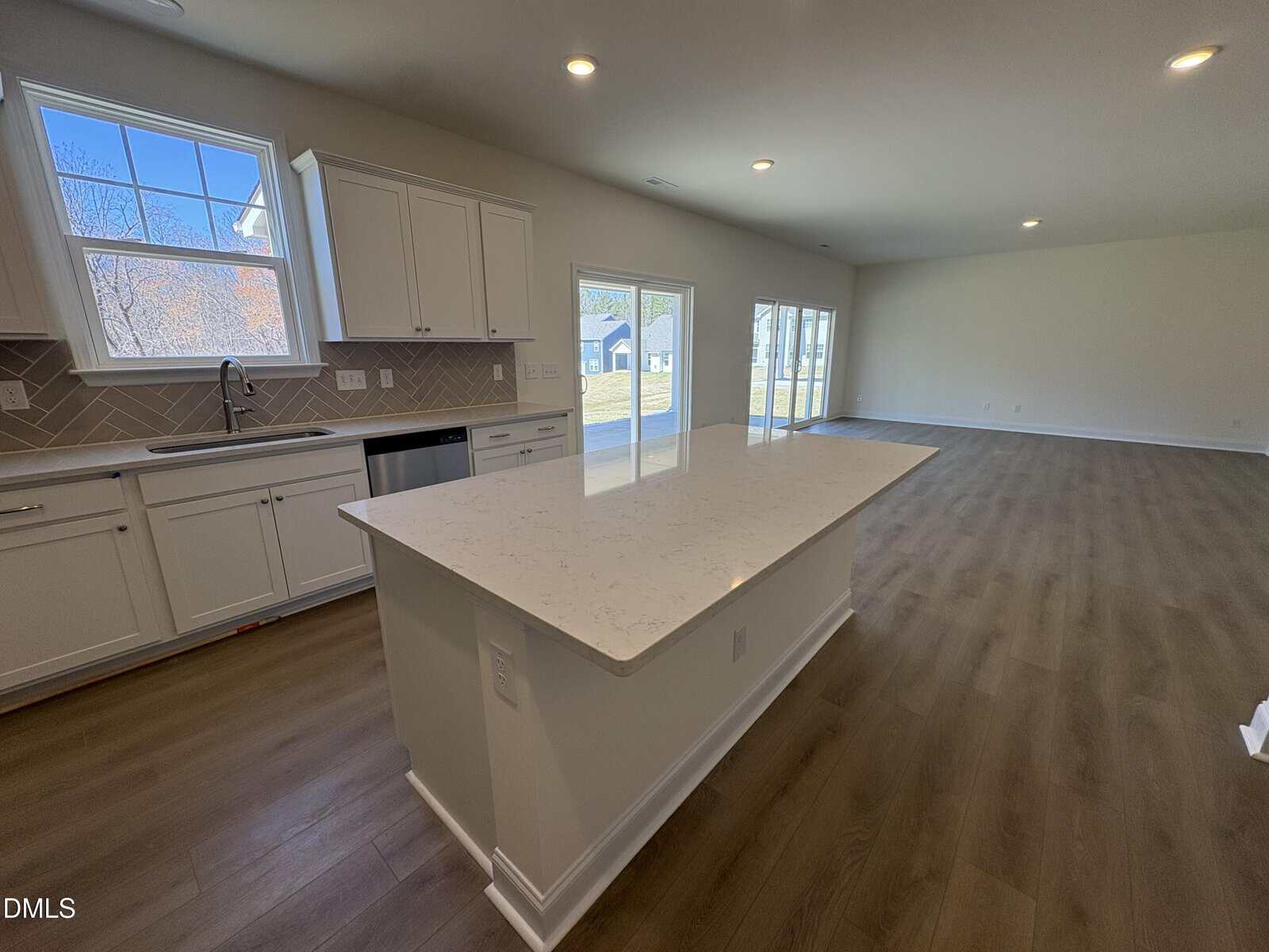 Spacious kitchen with white cabinets, a large island, and wood flooring, open to a bright living area with large windows and sliding glass doors to a backyard.