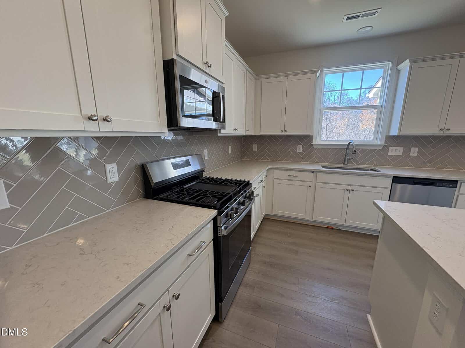 Modern kitchen with white cabinets, stainless steel appliances, gas stove, light countertops, tile backsplash, wood floors, and a window above the sink letting in natural light.