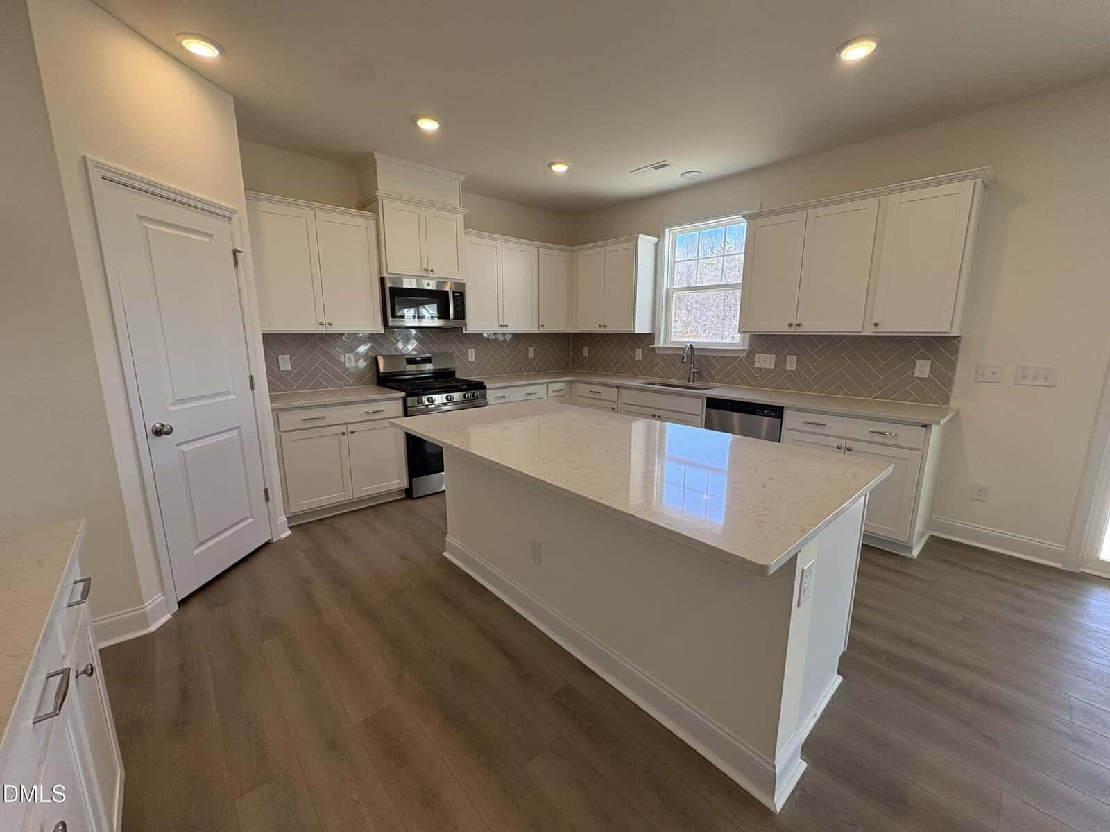 Modern kitchen with white cabinets, stainless steel appliances, an island with a quartz countertop, tile backsplash, wood flooring, and recessed lighting. A window above the sink lets in natural light.