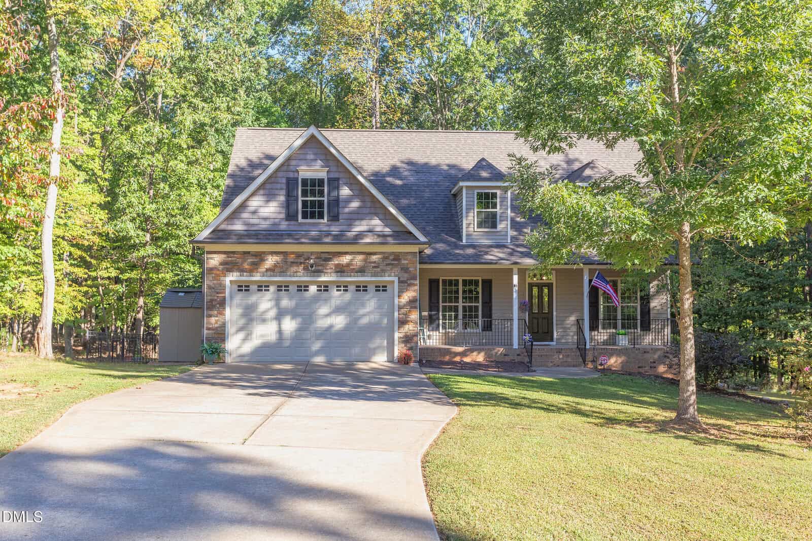 A two-story brick and siding house with a covered porch, attached two-car garage, and a concrete driveway, set among trees with a well-maintained lawn and a shed in the background.