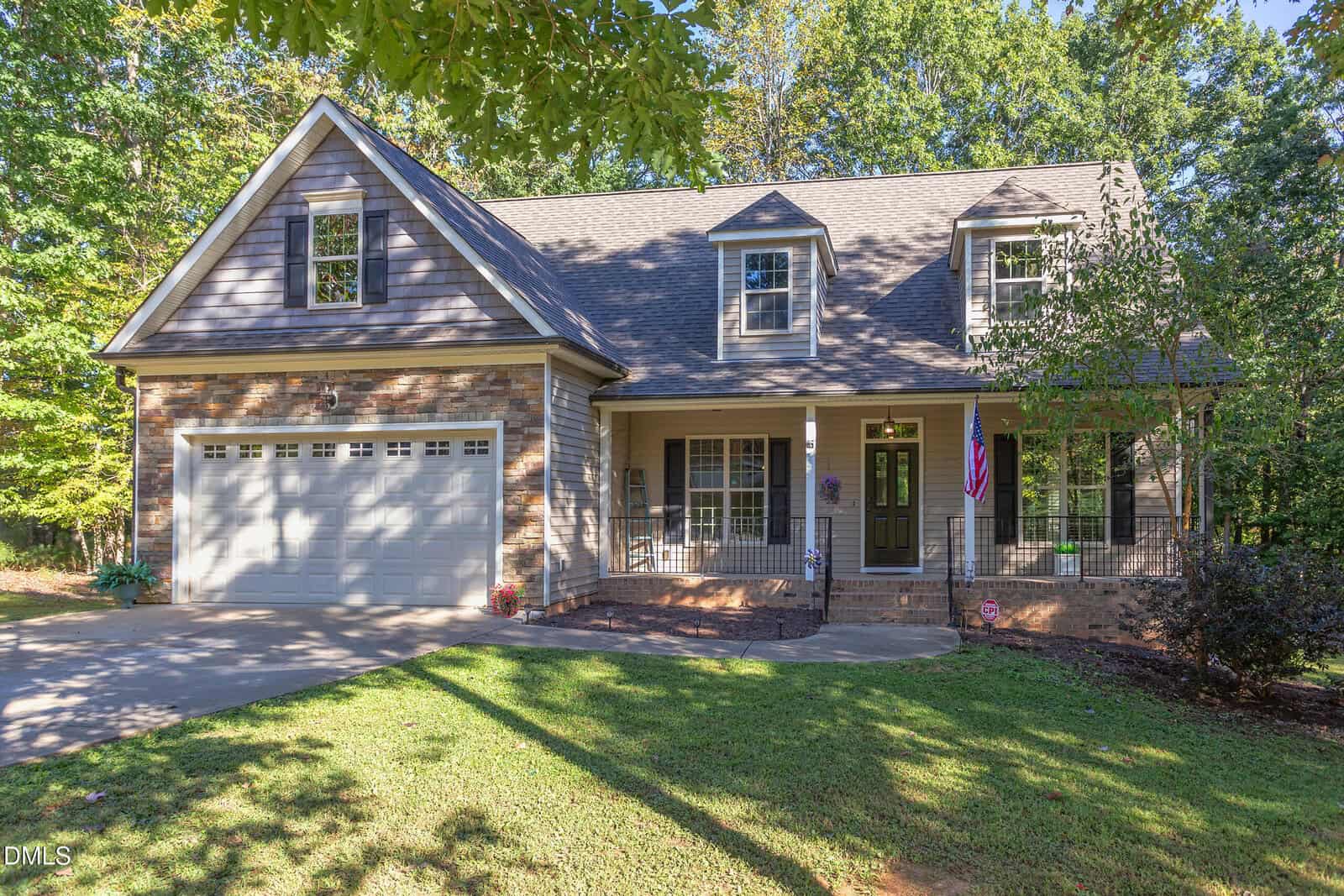 A two-story suburban house on 3627 Pine Needles Drive in Wake Forest with brick and siding exterior, an attached double garage, dormer windows, a covered front porch with an American flag, surrounded by trees and a front lawn.
