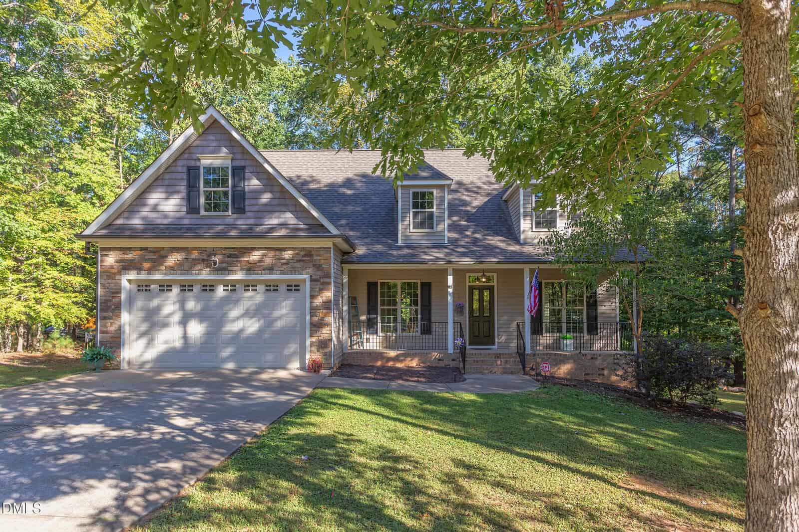 A two-story suburban house with a brick and siding exterior, an attached two-car garage, covered front porch, and a sloped driveway, set among trees with a well-maintained lawn.