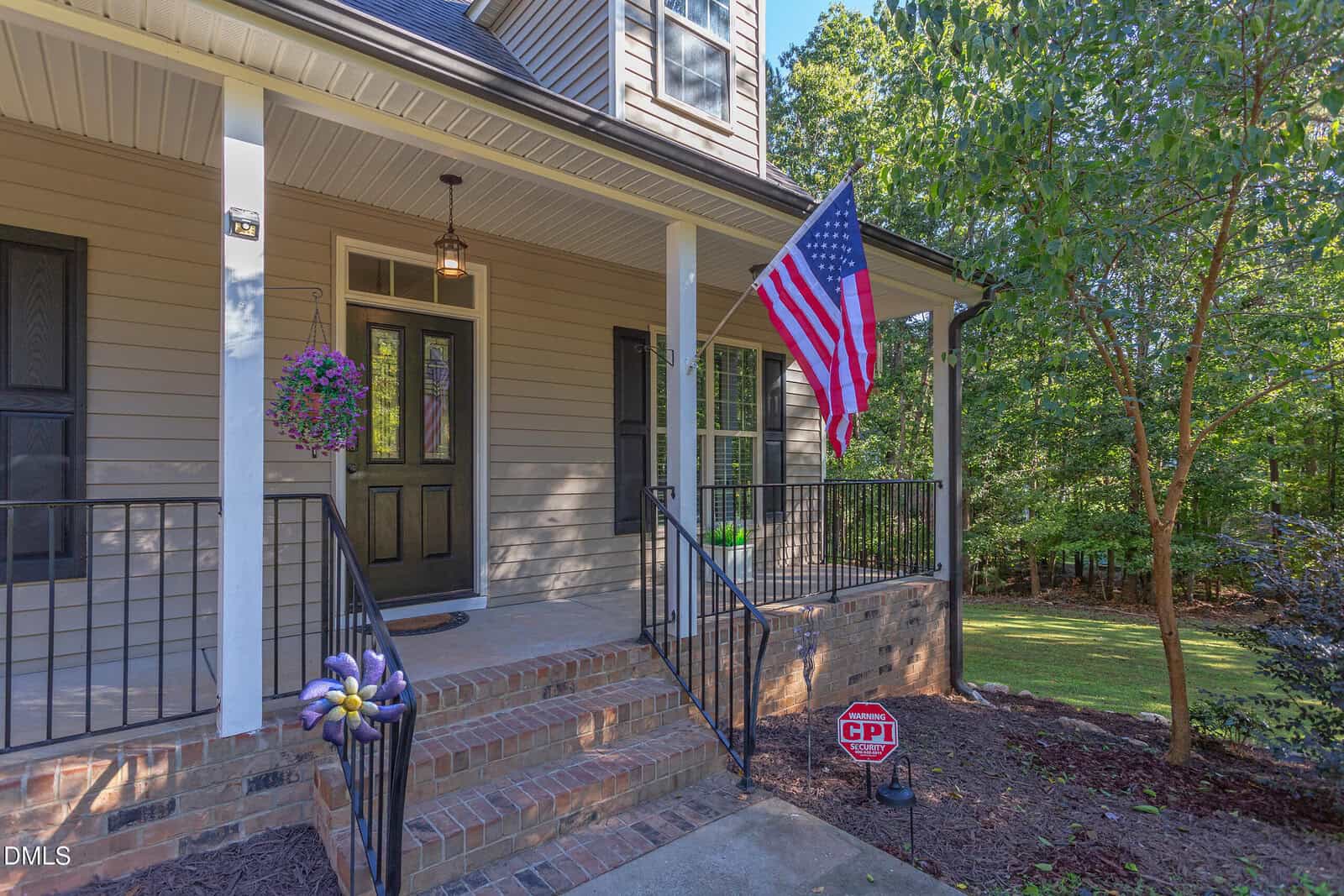 A front porch of a house with beige siding, black railing, brick steps, a hanging flower basket, an American flag, a CPI security sign, and a green yard with trees in the background.