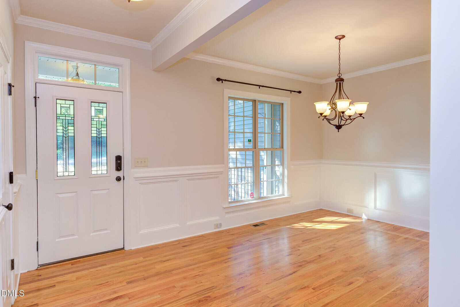 A bright, empty room with light hardwood floors, cream-colored walls with white wainscoting, a white front door with decorative glass, a large window, and a black metal chandelier.