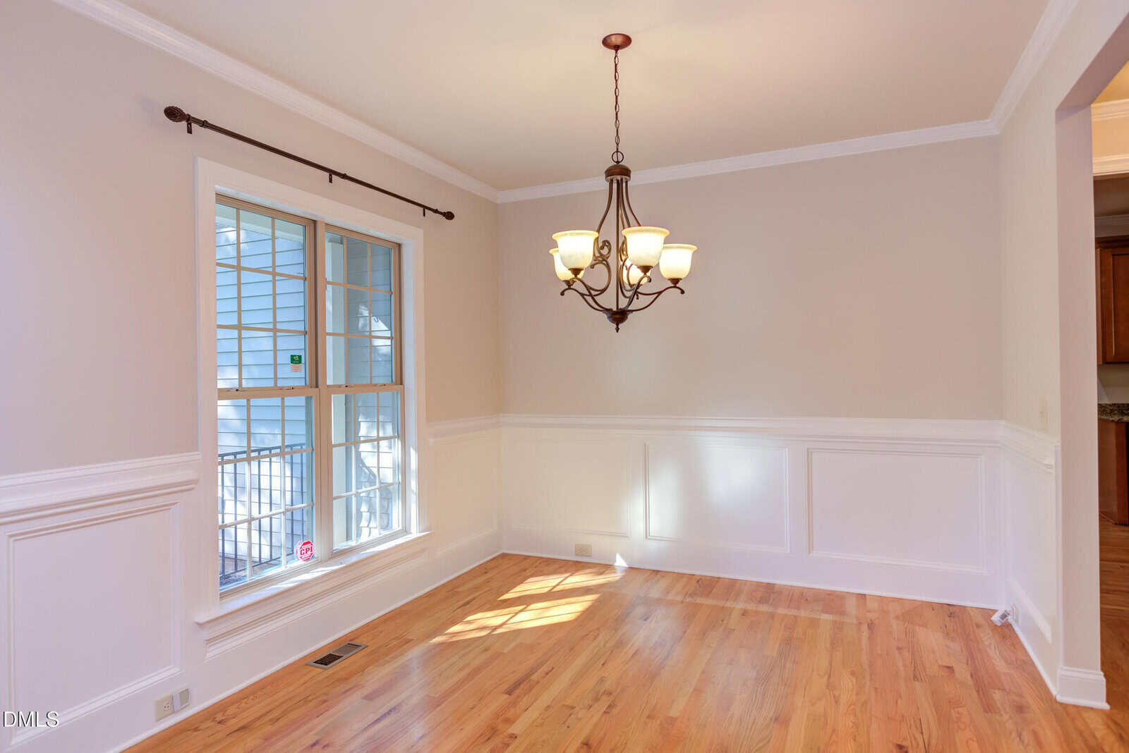 A bright dining room with light hardwood floors, beige walls with white wainscoting, a large window with grid panes, and a bronze chandelier hanging from the ceiling.