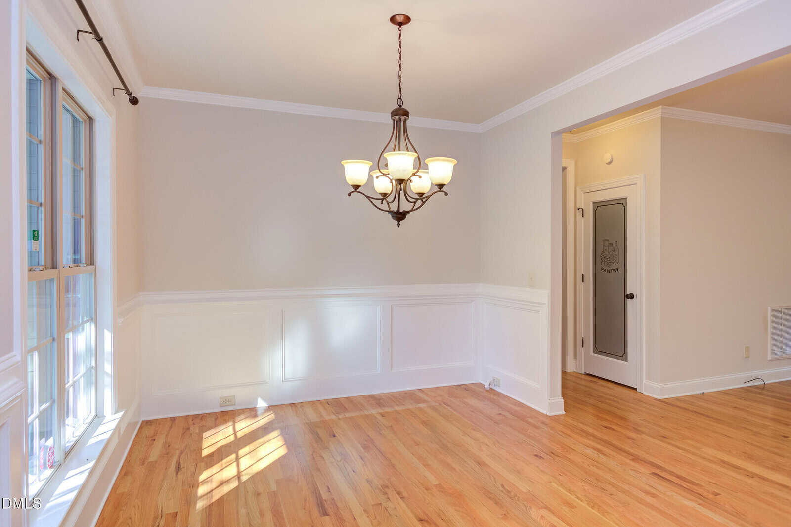 A dining room with hardwood floors, white wainscoting, beige walls, a window on the left, a six-light chandelier, and a door with frosted glass on the right side.