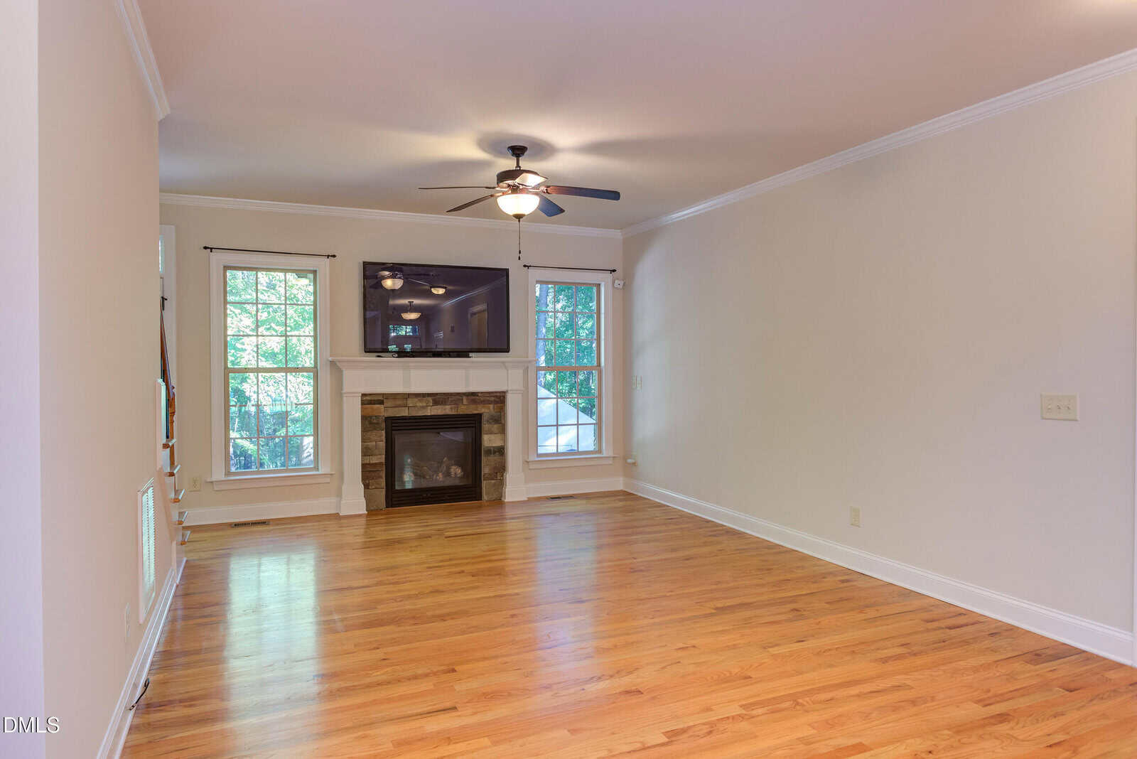 A living room with hardwood floors, a ceiling fan with a light, a fireplace with a mantel, a TV mounted above the fireplace, and three large windows letting in natural light. The walls are painted light beige.
