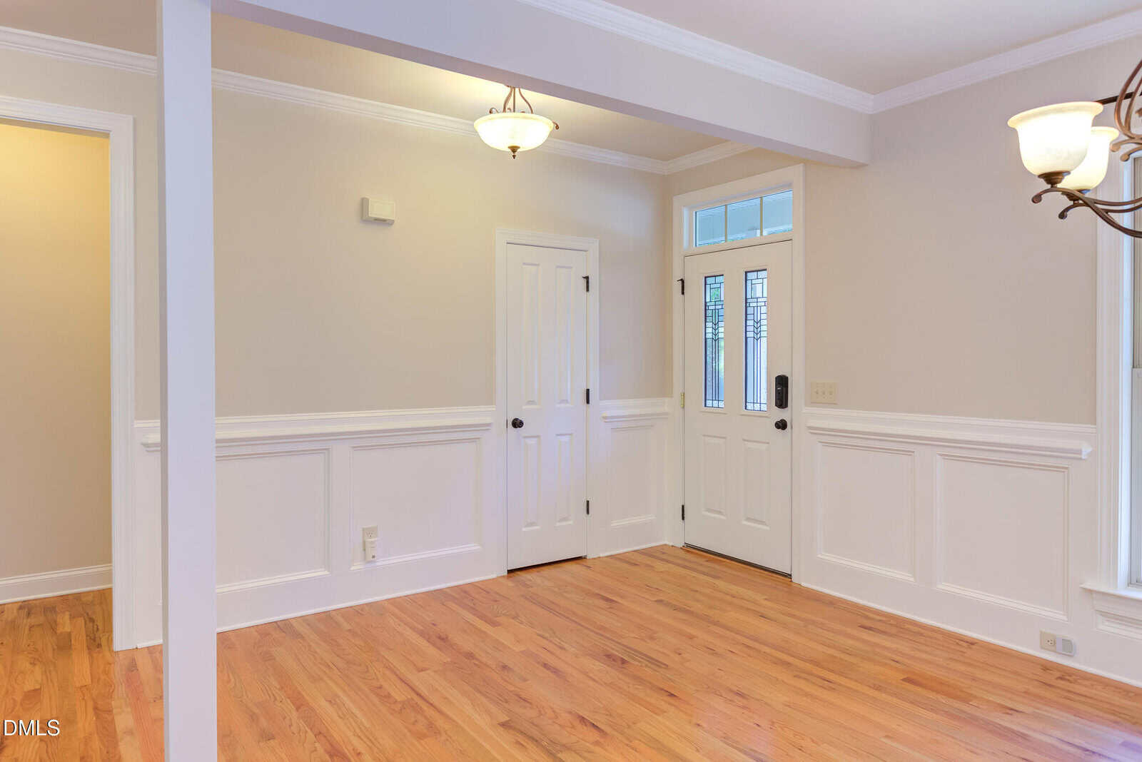 A room with light-colored walls, hardwood floors, white trim, and wainscoting. There are two doors, a front door with glass panes, and a ceiling light fixture. A second light fixture hangs partially in view.