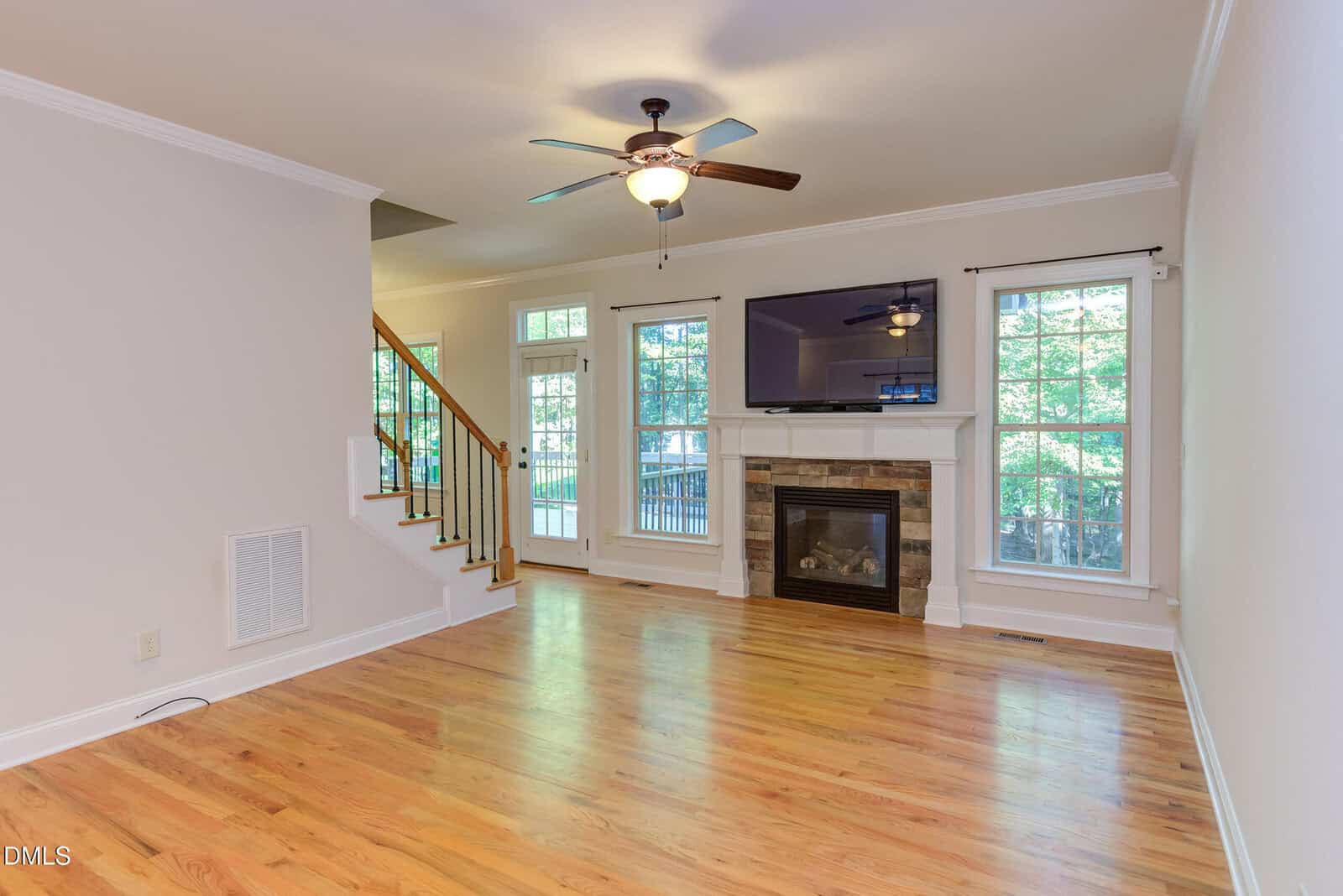 Bright living room with hardwood floors, ceiling fan, fireplace with stone trim, mounted TV, large windows, and a staircase with wooden railing leading upstairs. Natural light fills the space, giving an open, airy feel.
