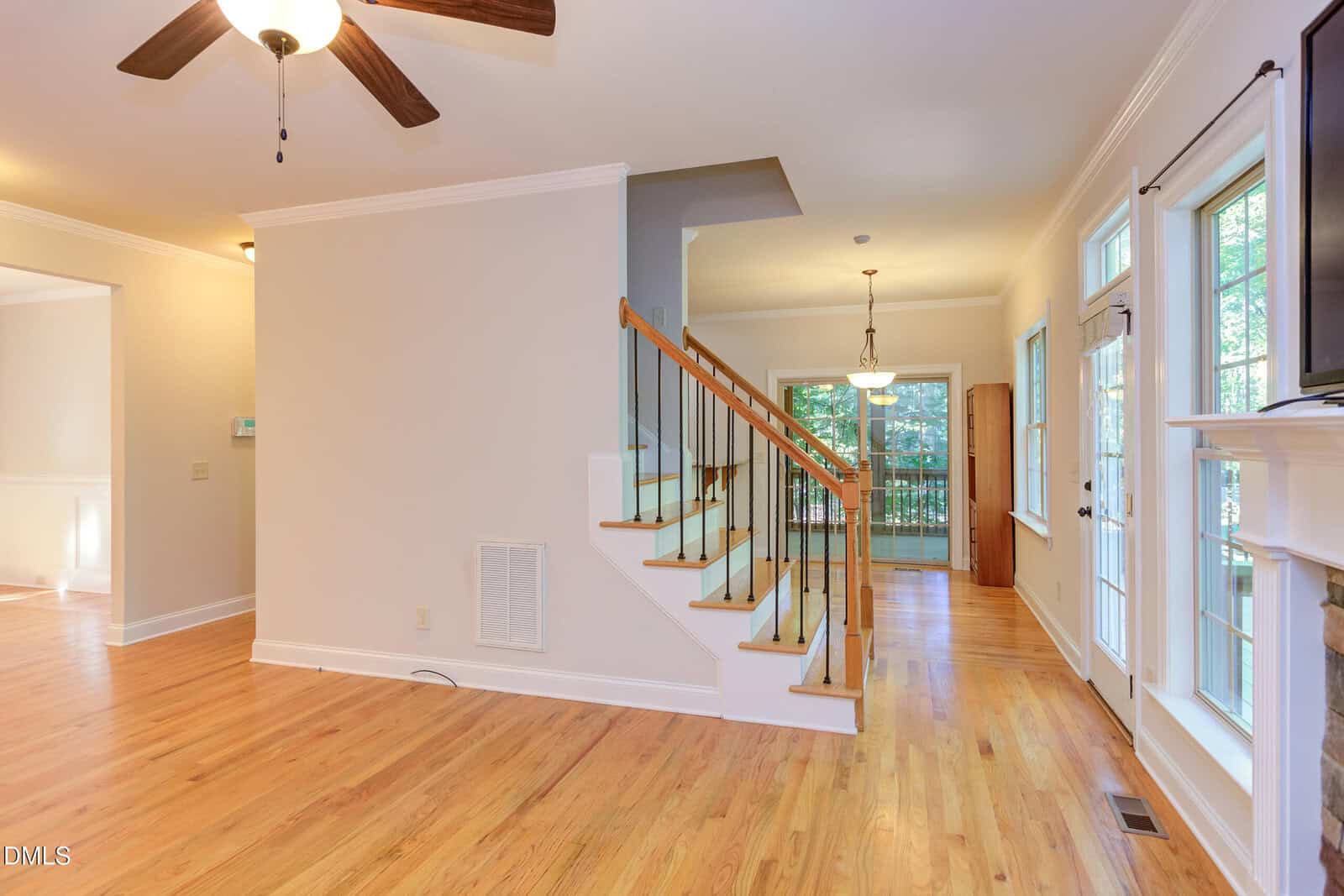 Bright living area with hardwood floors, a staircase with wood and iron railing, ceiling fan, and large windows. Double doors lead outside; dining space visible near the back with a hanging light fixture.