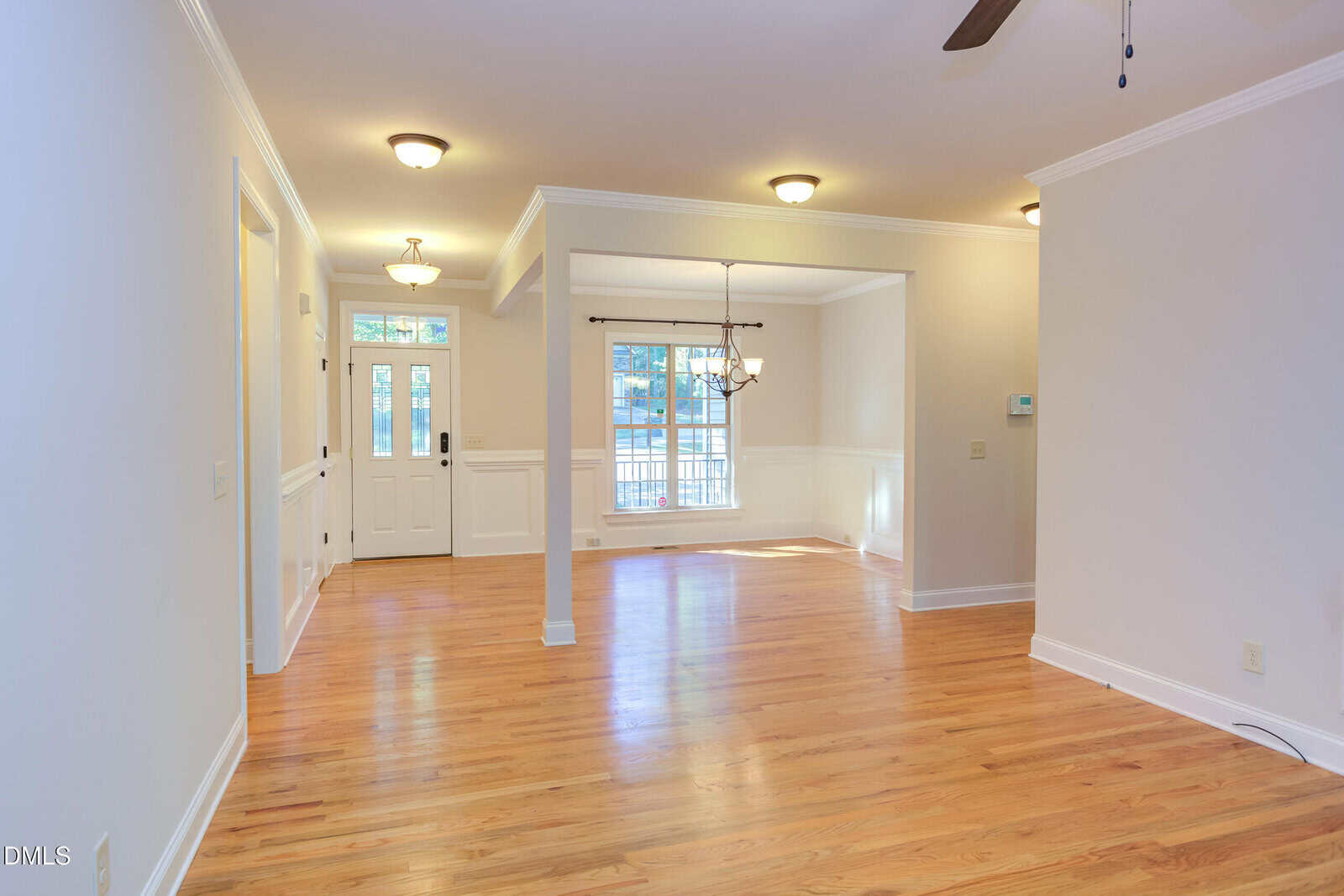 Bright, empty living room with hardwood floors, white walls, ceiling lights, crown molding, and a windowed front door. Large windows and a hanging light fixture illuminate the dining area.