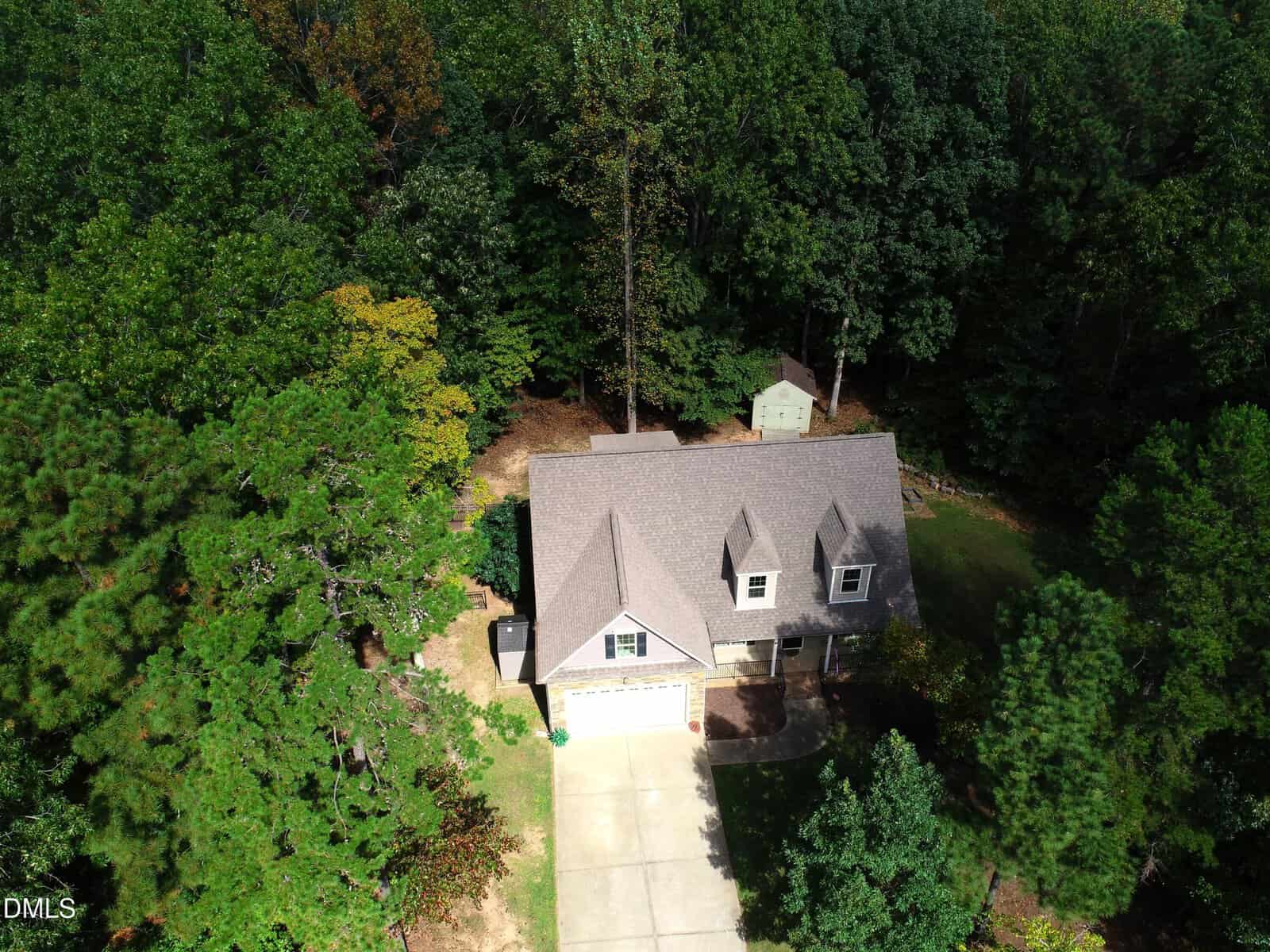 Aerial view of a two-story house with a gray roof and attached garage, surrounded by dense trees. A driveway leads to the garage, and a small shed is visible in the backyard near the edge of the woods.