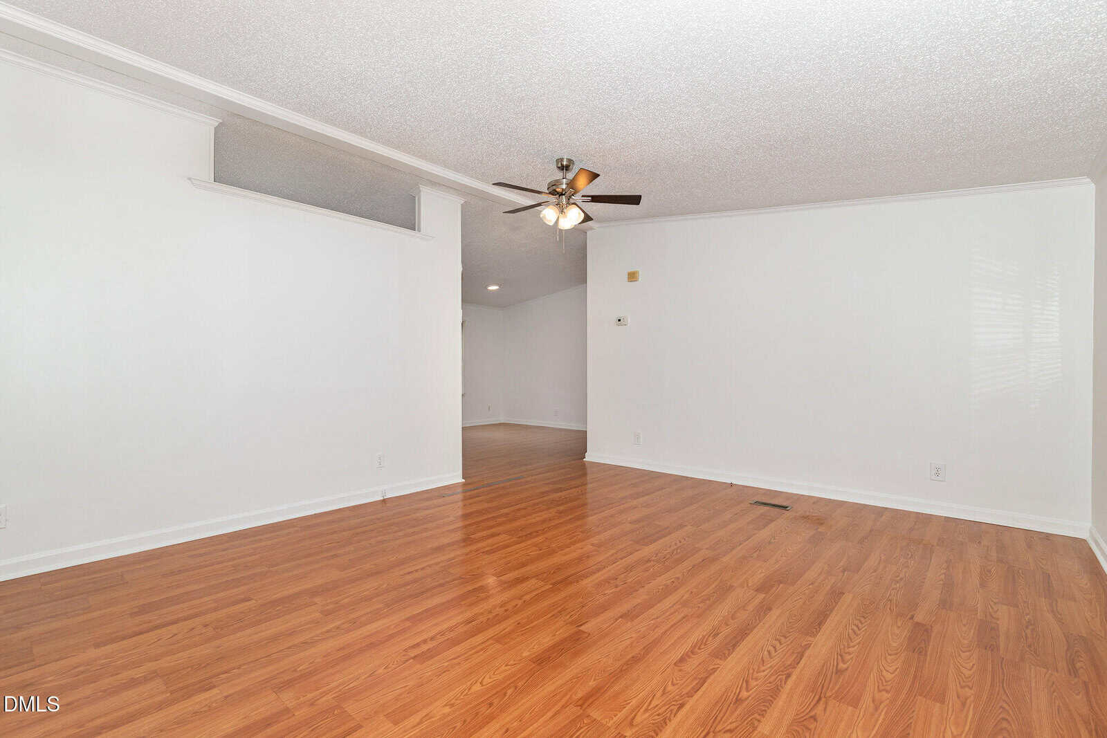 A room with white walls, wood laminate flooring, a ceiling fan with lights, and a partial interior window above one wall. There’s a small vent on the floor and a glimpse into another room in the background.