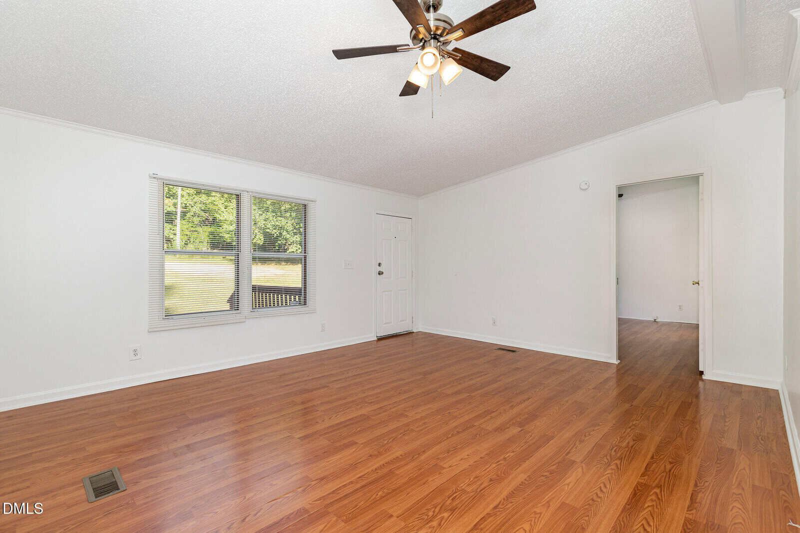 A bright, empty room with wood flooring, white walls, a ceiling fan with lights, two windows, and a white door. There is an open doorway leading to another room with similar flooring.