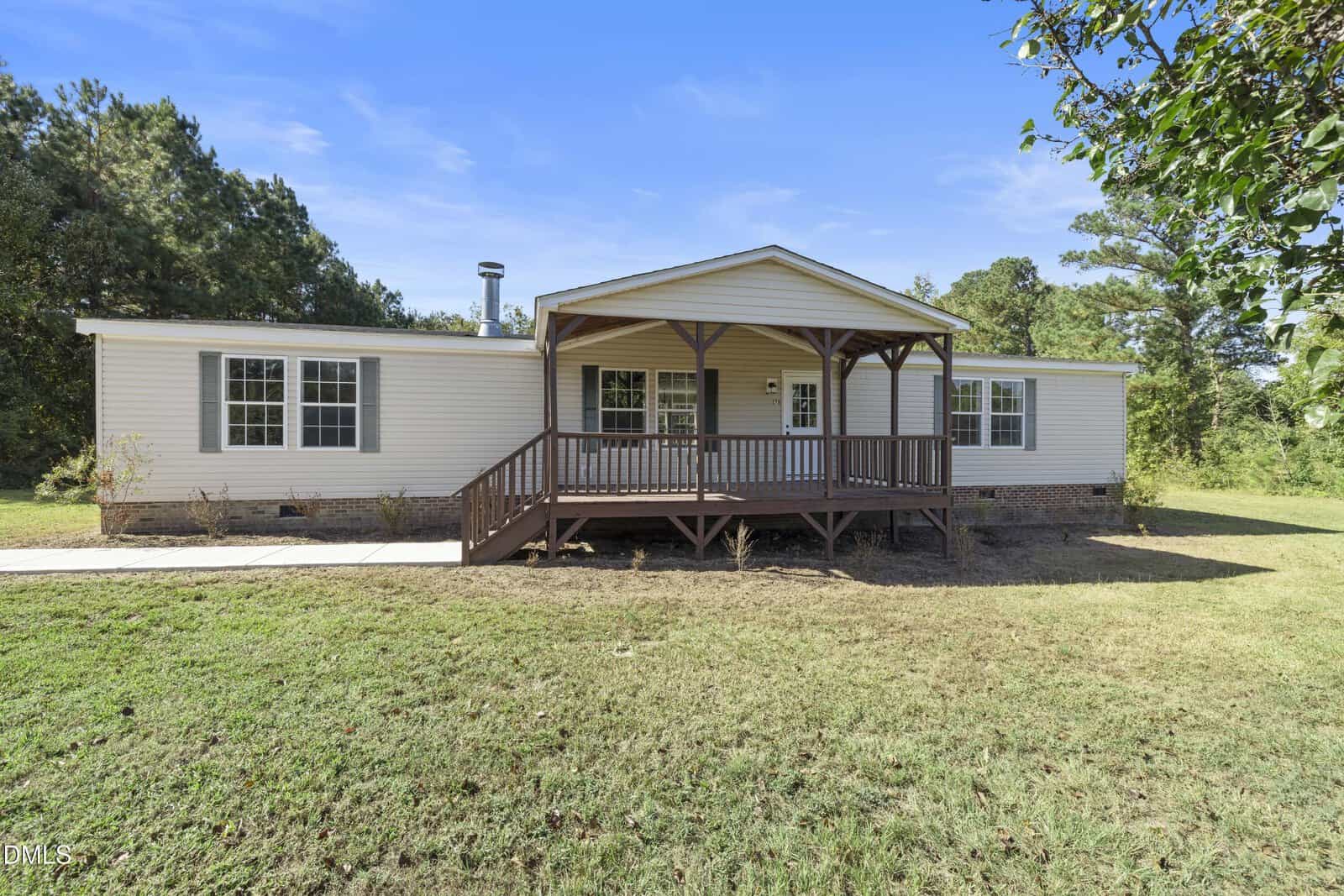 A single-story manufactured home with beige siding and a covered wooden front porch, set on a grassy lawn with trees in the background under a clear blue sky.
