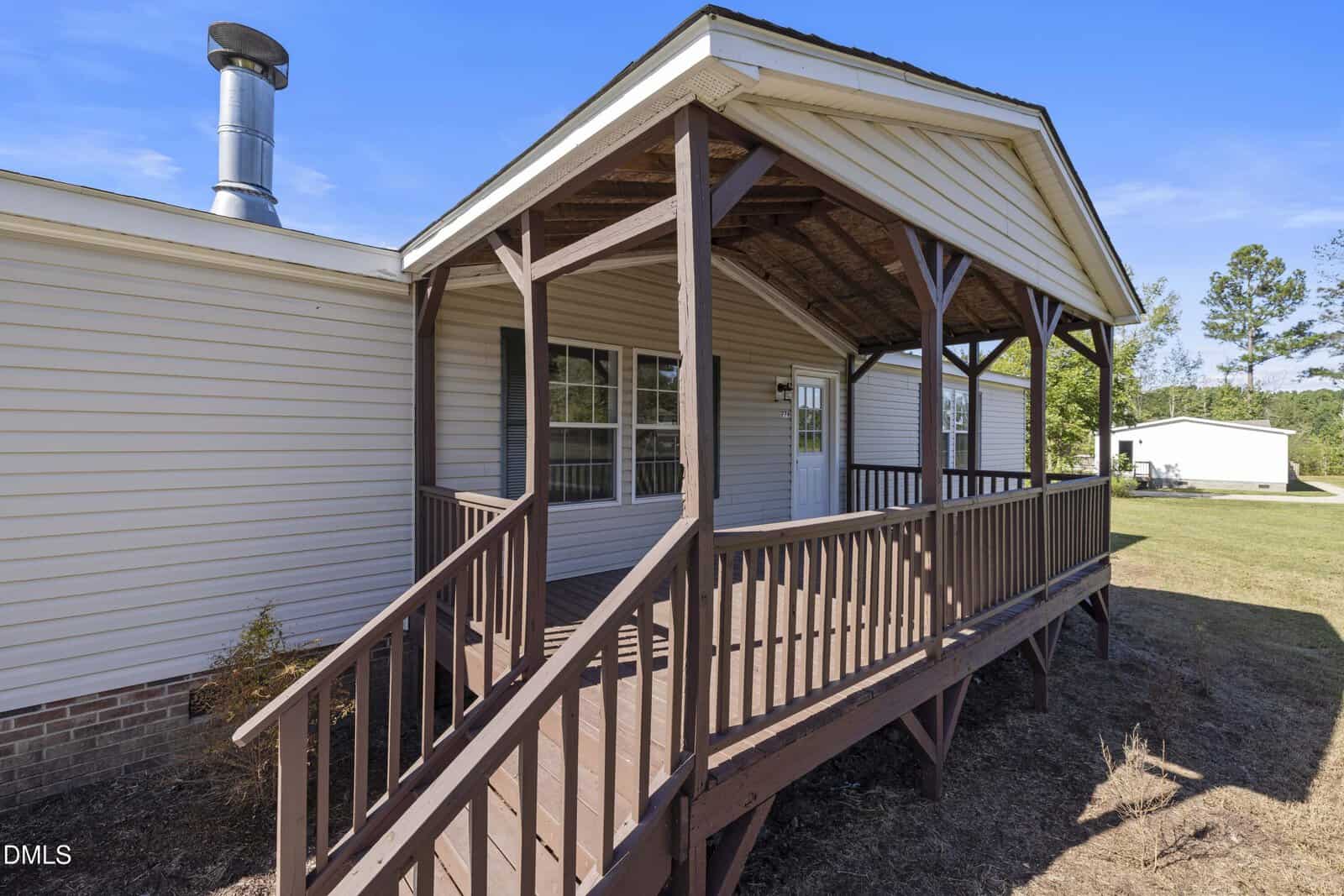 A brown wooden ramp and porch lead to the white door of a single-story beige house with siding. The area around the porch is bare soil, and a detached white building is visible in the background.