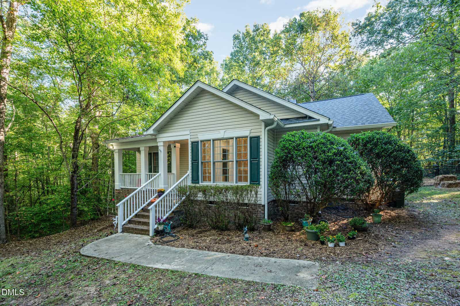 A small, single-story house with green shutters, a covered front porch, white steps, and several potted plants. The house is surrounded by trees and sits on a dirt and concrete path.