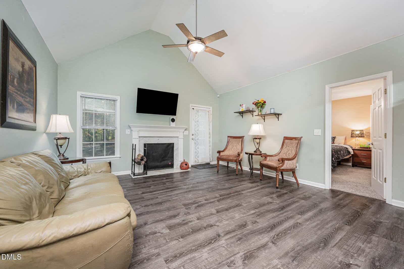 Living room with light green walls, wood floor, beige sofa, two chairs, a ceiling fan, wall-mounted TV, fireplace, two lamps, and a small door. An open doorway leads to a bedroom with a bed and nightstand.