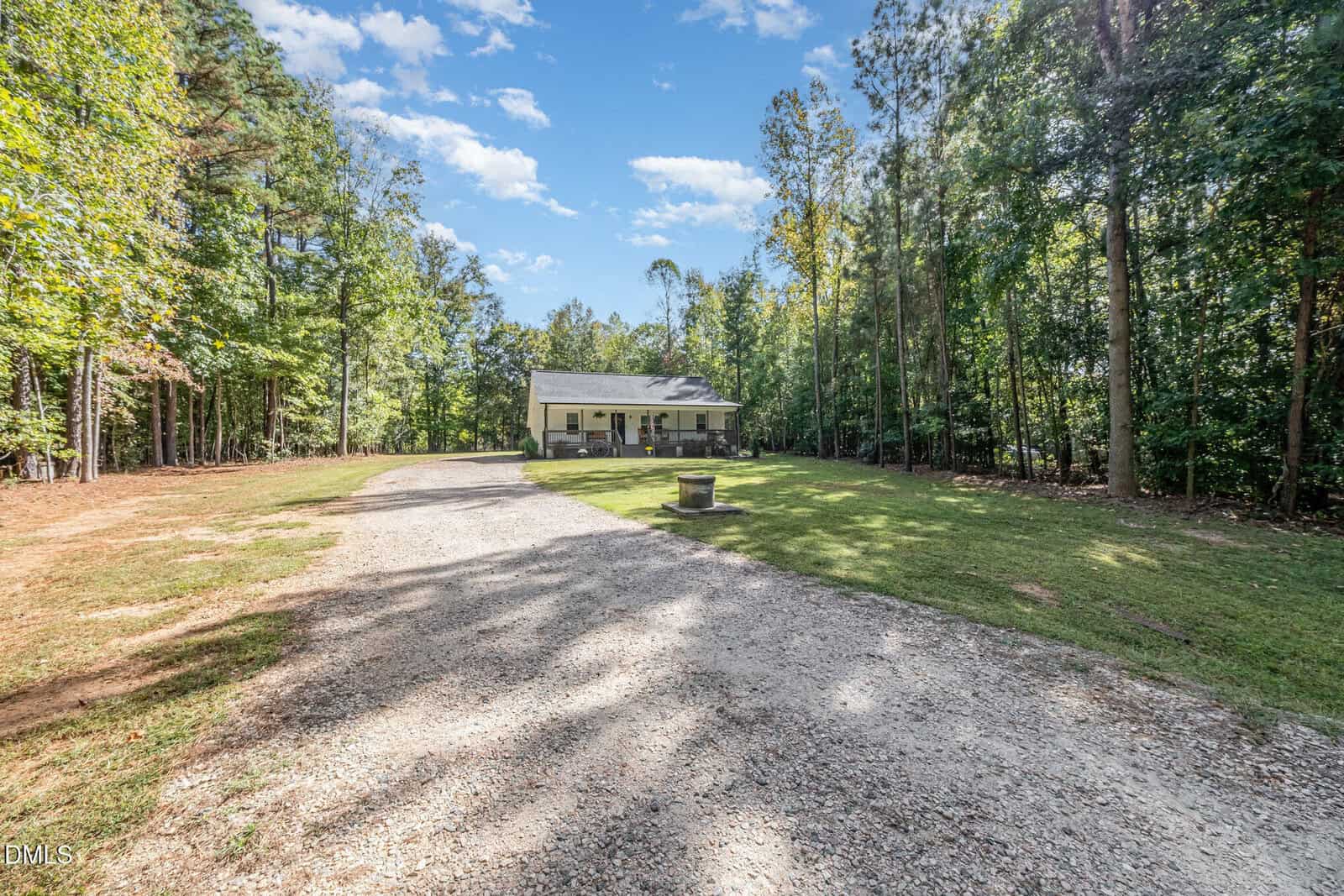 A white house with a covered front porch sits at the end of a long gravel driveway, surrounded by tall trees and green lawn under a blue sky with scattered clouds.