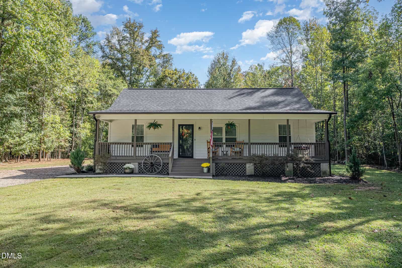 A charming one-story white house at 105 Hines Court, Youngsville, featuring a covered front porch, black roof, and inviting steps leading to the entrance. The home is surrounded by a lush green lawn and mature trees and boasts classic wagon wheel décor on the spacious porch.