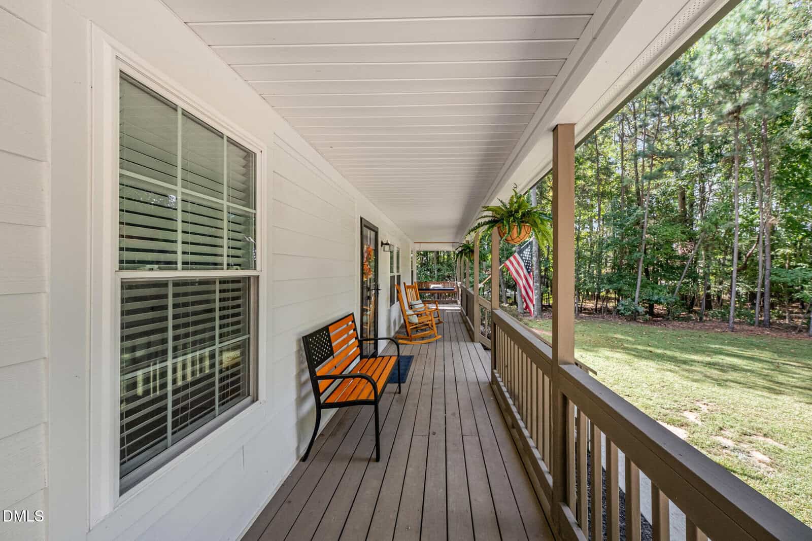 A covered front porch features a black bench with orange cushions, rocking chairs, hanging fern, American flag, and a railing. The porch overlooks a green lawn and is bordered by tall trees.