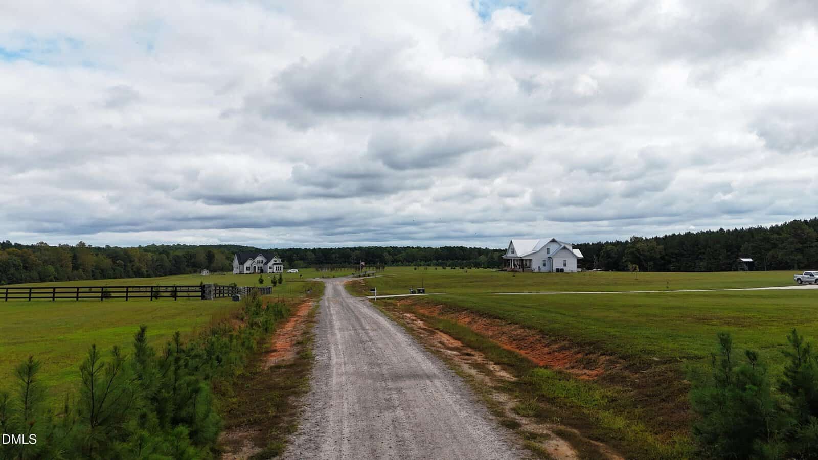 A gravel road runs through a large grassy field with scattered trees, leading to a white house and other buildings in the distance under a cloudy sky.