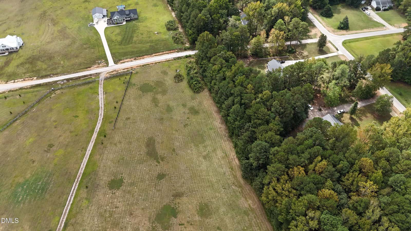 Aerial view of a rural area with open grassy fields on the left, a line of trees separating the fields from a residential area with houses and driveways on the right. A road runs horizontally near the top of the image.