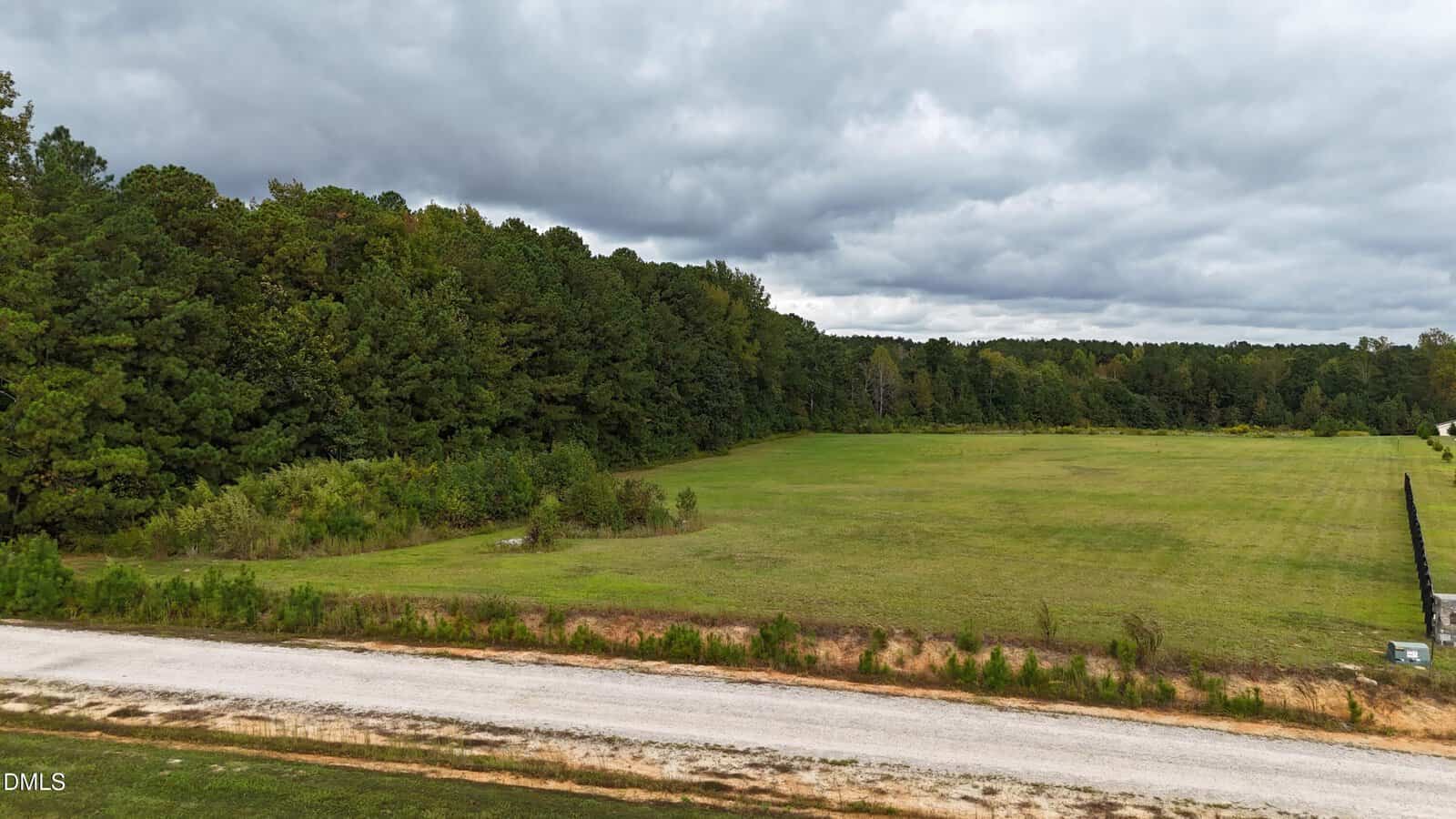 A grassy, open field bordered by dense trees under a cloudy sky, with a narrow gravel road running along the front edge of the field.