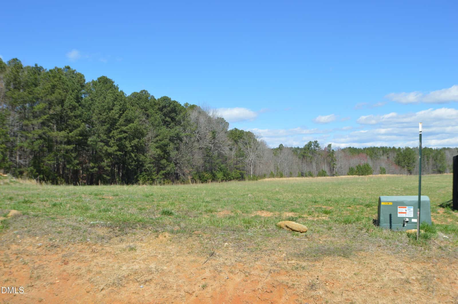 Open grassy field with a patch of exposed dirt in the foreground, bordered by dense trees under a clear blue sky. A green utility box and a white pole are visible on the right side.