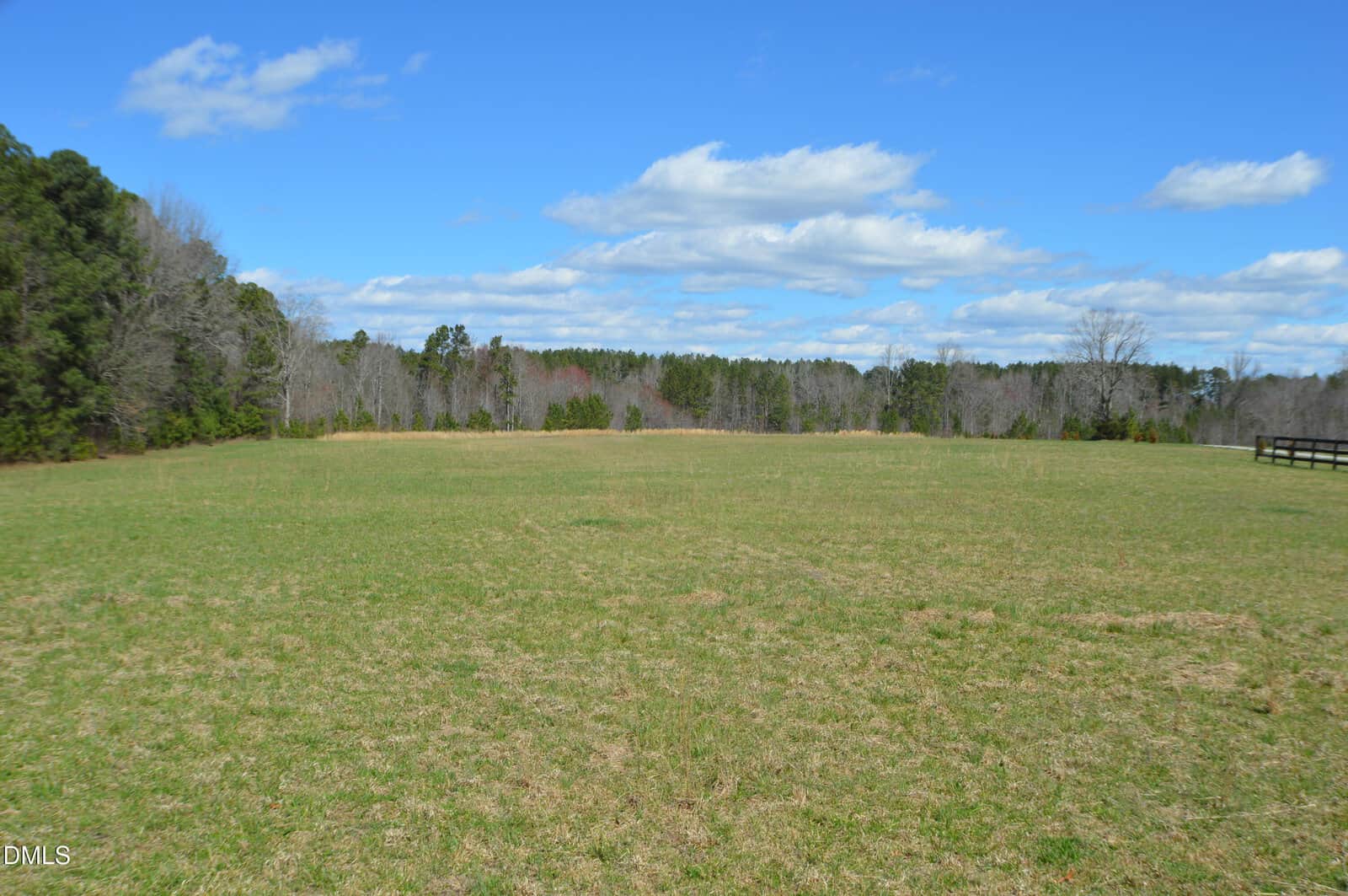 A large open grassy field under a blue sky with scattered white clouds. Trees line the background, and part of a wooden fence is visible on the right side of the image.