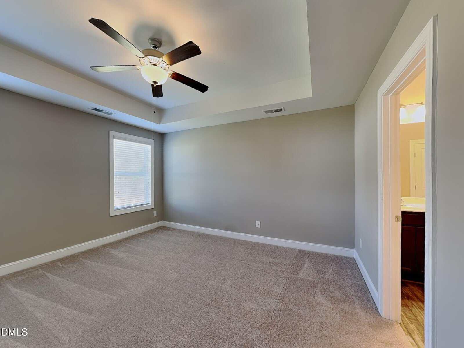 Empty bedroom with beige carpet, light gray walls, and a tray ceiling with a ceiling fan and light. A window provides natural light, and an open doorway leads to a bathroom with visible sink and mirror.
