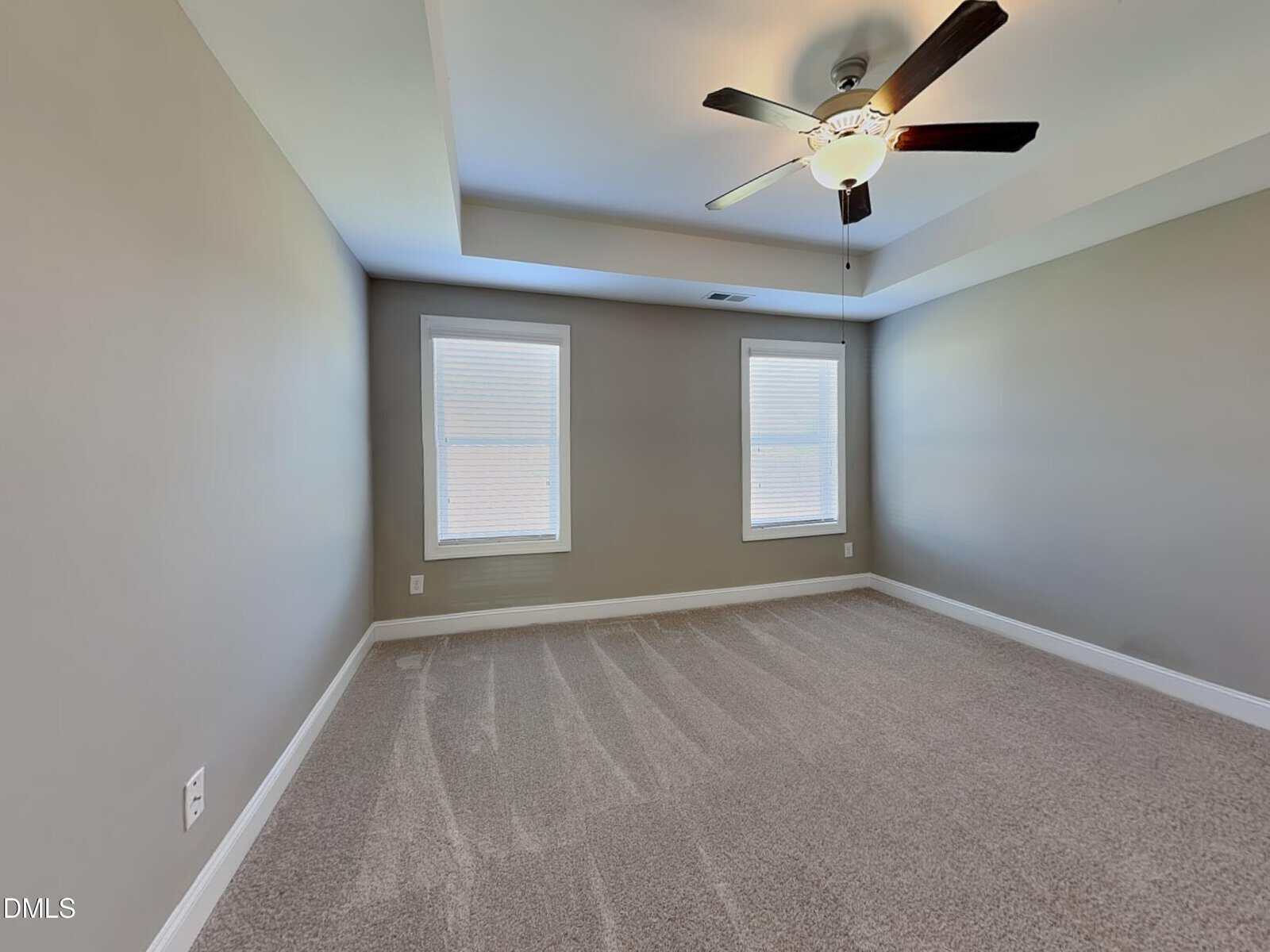 A vacant room with beige carpet, taupe walls, two windows with closed blinds, a white ceiling with a ceiling fan and light fixture, and white baseboards.