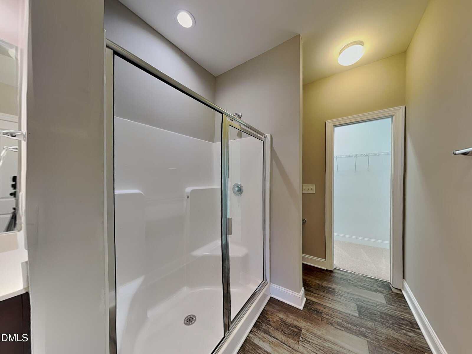 Modern bathroom with a glass shower enclosure, wood-look flooring, beige walls, recessed lighting, and a view into an adjacent walk-in closet with built-in shelving.
