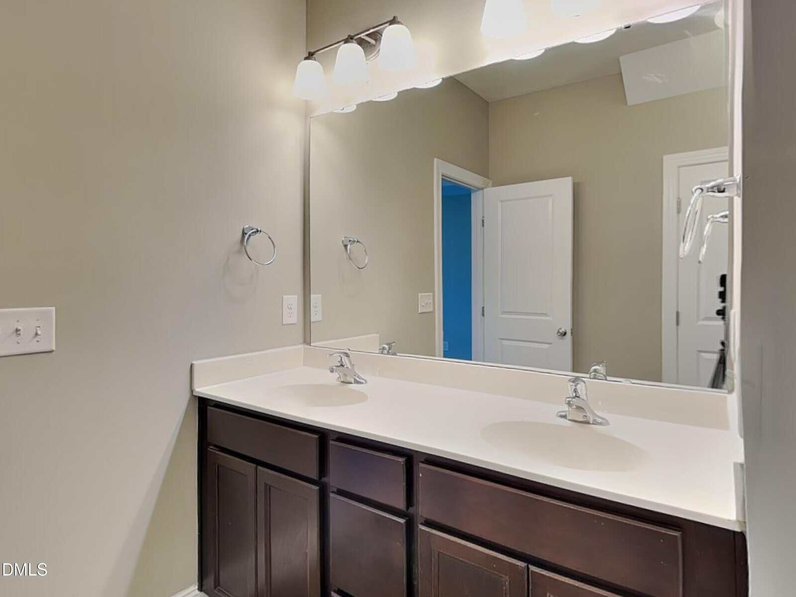 A bathroom with a double sink vanity featuring dark cabinets, a large mirror, wall-mounted light fixtures, two faucets, beige walls, and a white door visible in the background.