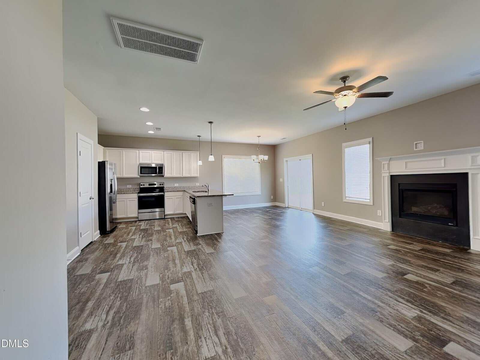 Open concept kitchen and living area with wood-look flooring, white cabinets, stainless steel appliances, central island, ceiling fan, fireplace, and multiple windows letting in natural light.