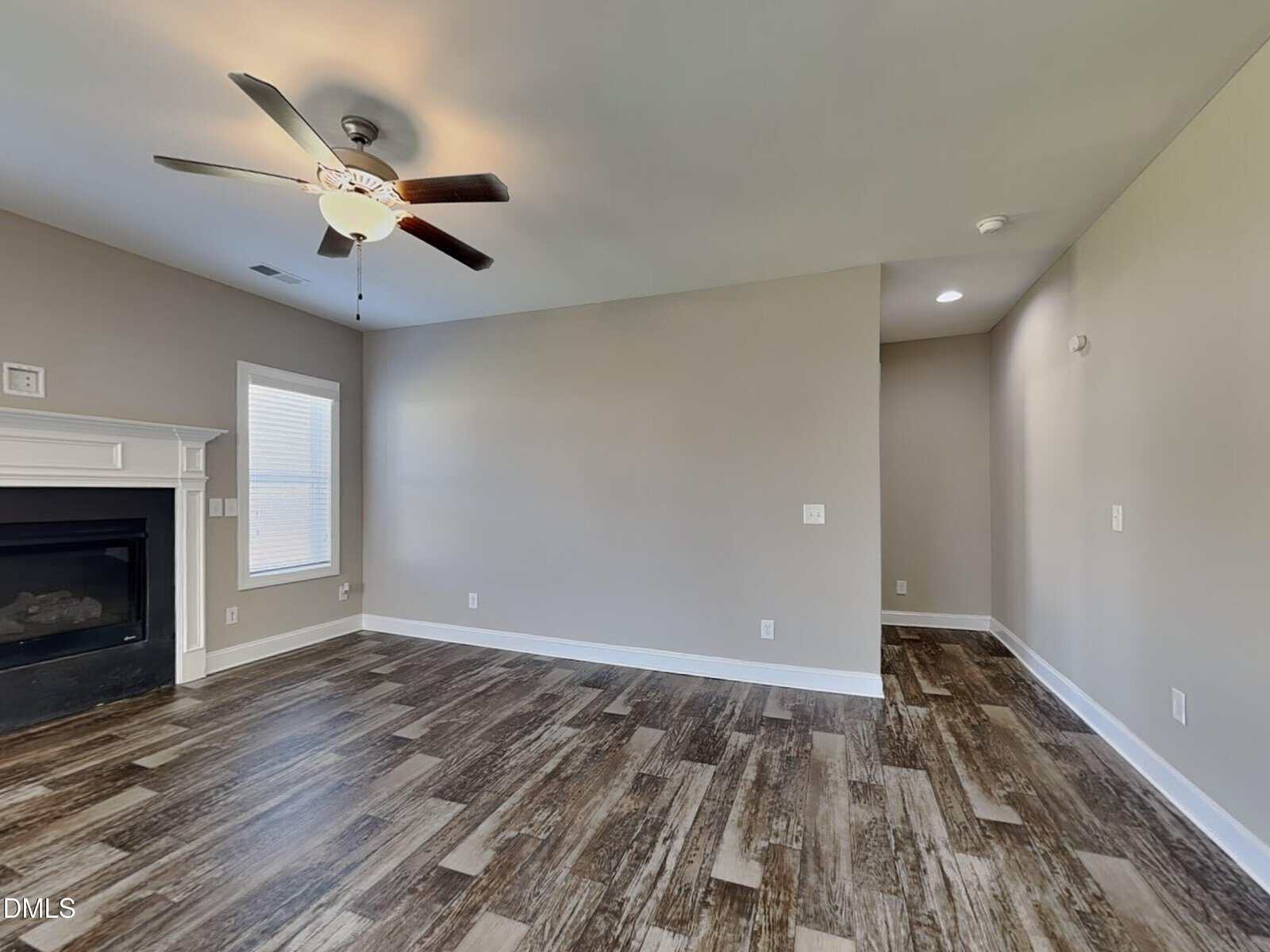 A room with wood-style flooring, beige walls, a ceiling fan with lights, a white fireplace on the left, and a window beside it. An alcove or hallway is visible to the right of the main area.