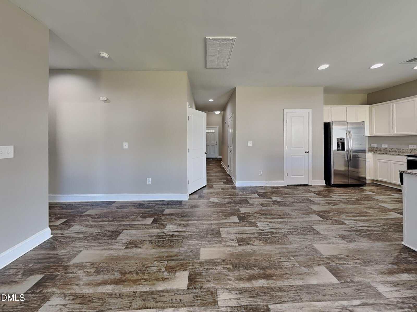 Open-concept kitchen and living area with beige walls, wood-patterned tile flooring, white cabinets, stainless steel appliances, and multiple doors leading to other rooms. Room is well-lit with ceiling lights.