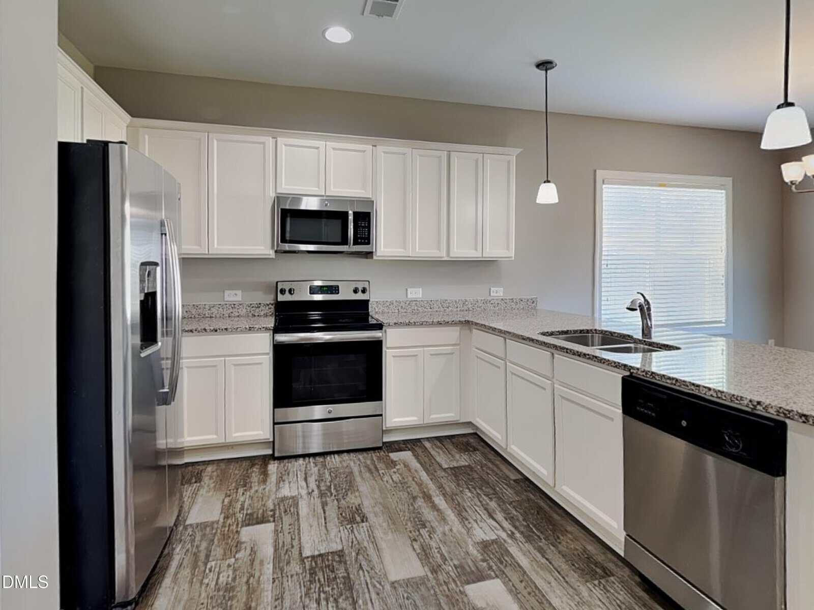 Modern kitchen with white cabinets, stainless steel appliances, granite countertops, double sink, wood-look flooring, and pendant lighting. A window with blinds is visible near the sink.