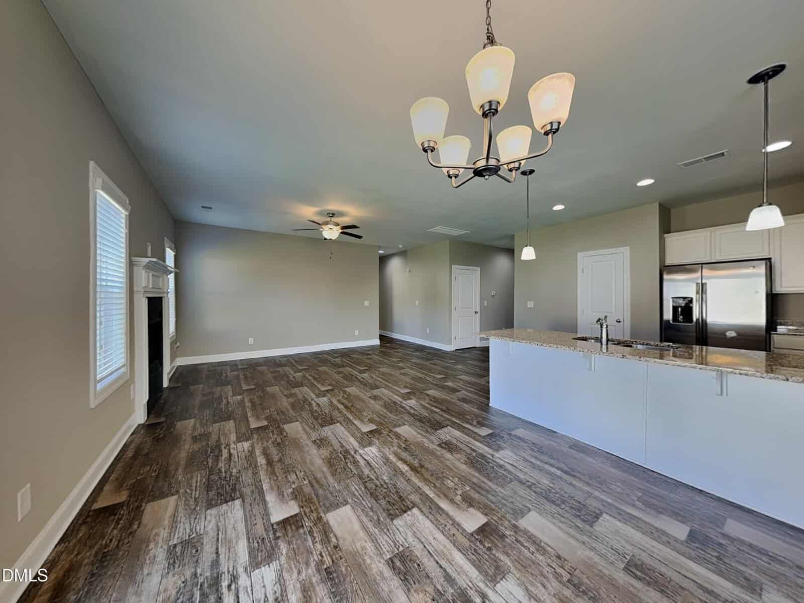 Open-concept kitchen and living area with wood-look tile flooring, light gray walls, ceiling fans, modern lighting fixtures, and large windows. The kitchen features a granite countertop and stainless steel appliances.