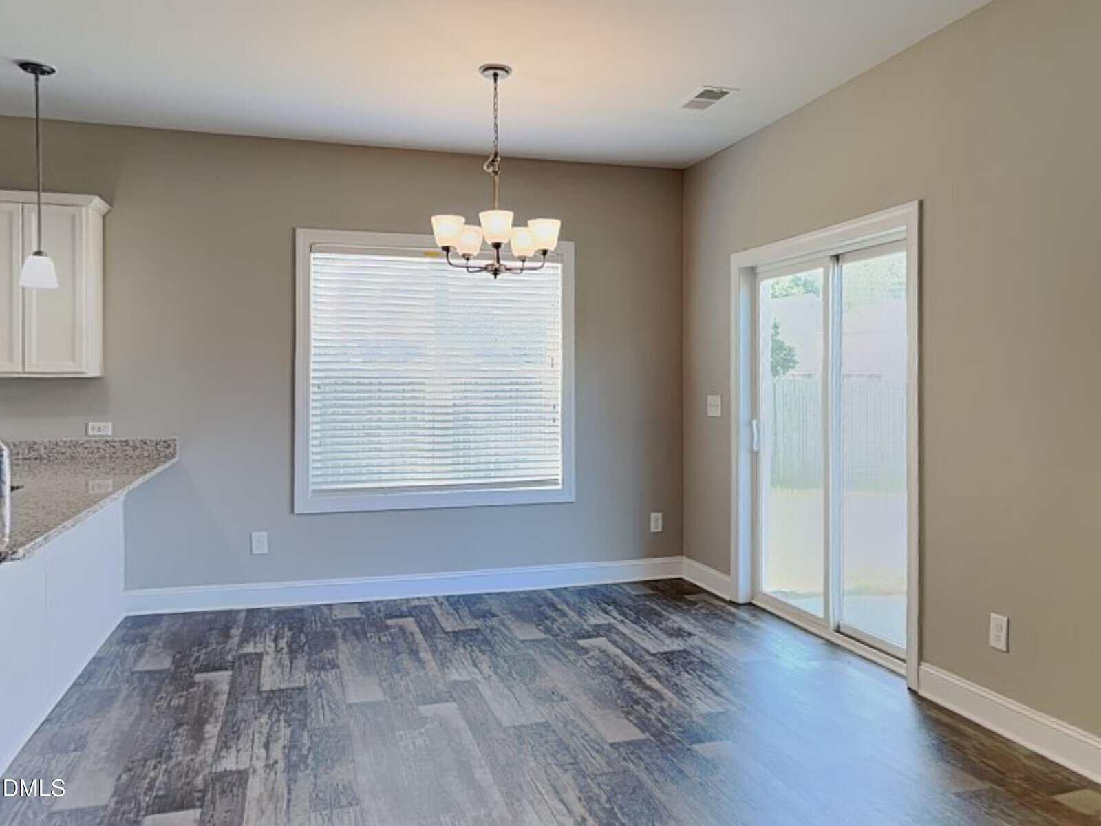 A dining area with dark wood flooring, light gray walls, a window with blinds, a ceiling light fixture, and sliding glass doors leading outside, adjacent to a kitchen countertop.