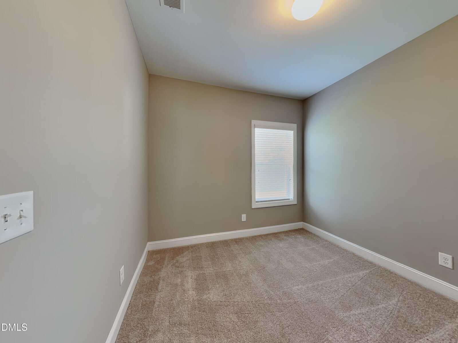 Empty room with beige carpet, light gray walls, a white electrical outlet, a ceiling light, and a single window with closed blinds letting in some natural light.