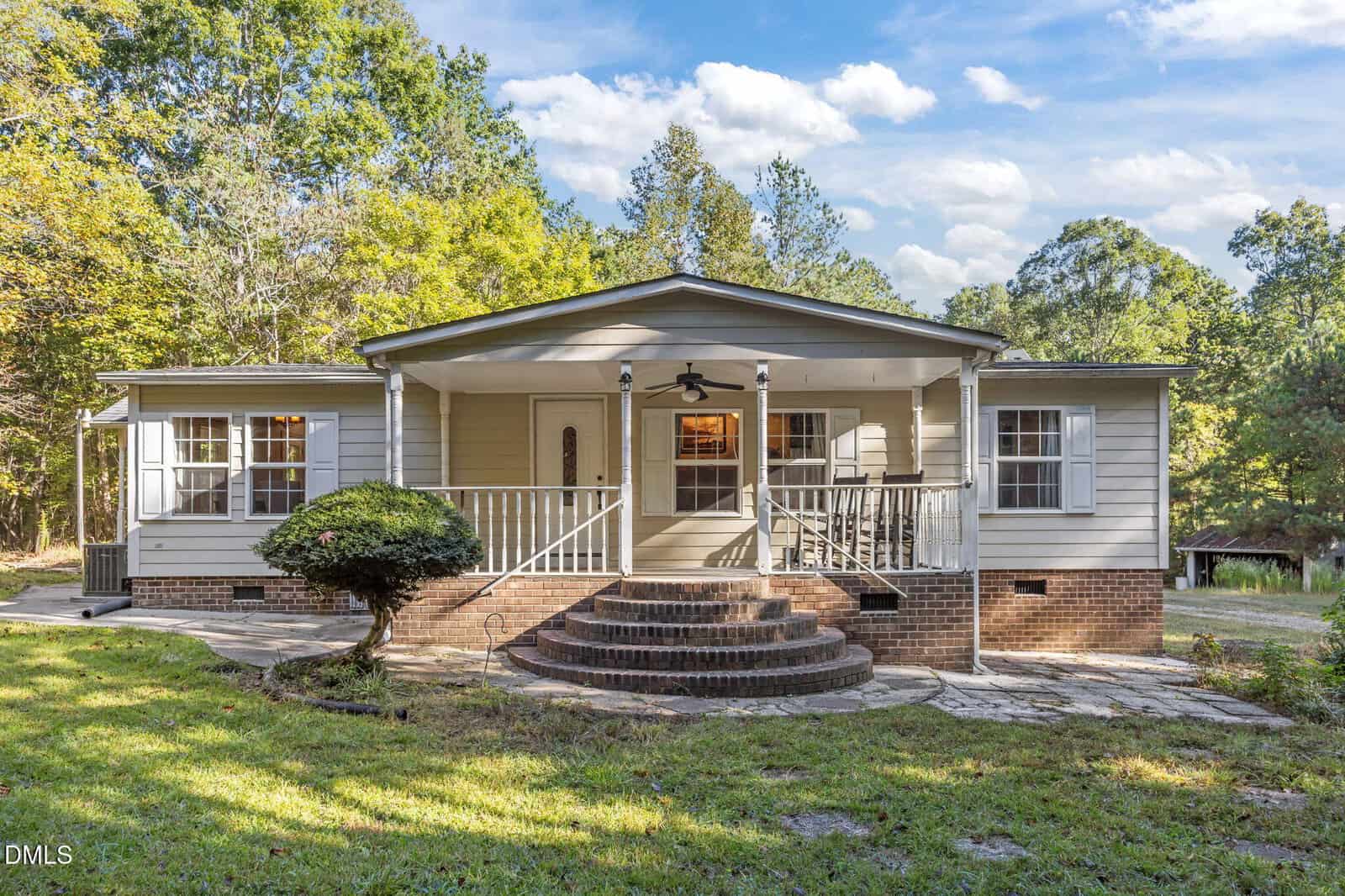 Single-story house at 328 N Raleigh Farms Road in Youngsville with light gray siding and a brick foundation, featuring a covered front porch with steps, white railings, and a ceiling fan. Surrounded by trees, this home welcomes you with a curved stone walkway leading to the entrance.
