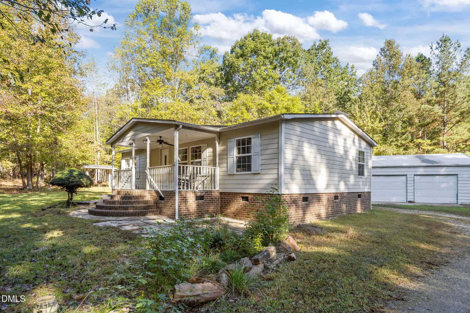 Single-story light gray house with a front porch, brick foundation, and steps leading up to the entrance, situated in a wooded area with trees and a gravel driveway. A white outbuilding is visible in the background.
