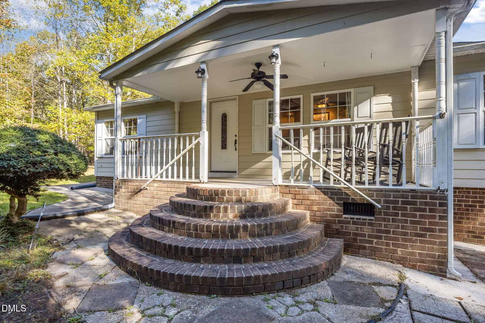 Front view of a house with beige siding, brick foundation, and curved brick steps leading to a covered porch with rocking chairs, a ceiling fan, and white columns. A trimmed bush is visible on the left.
