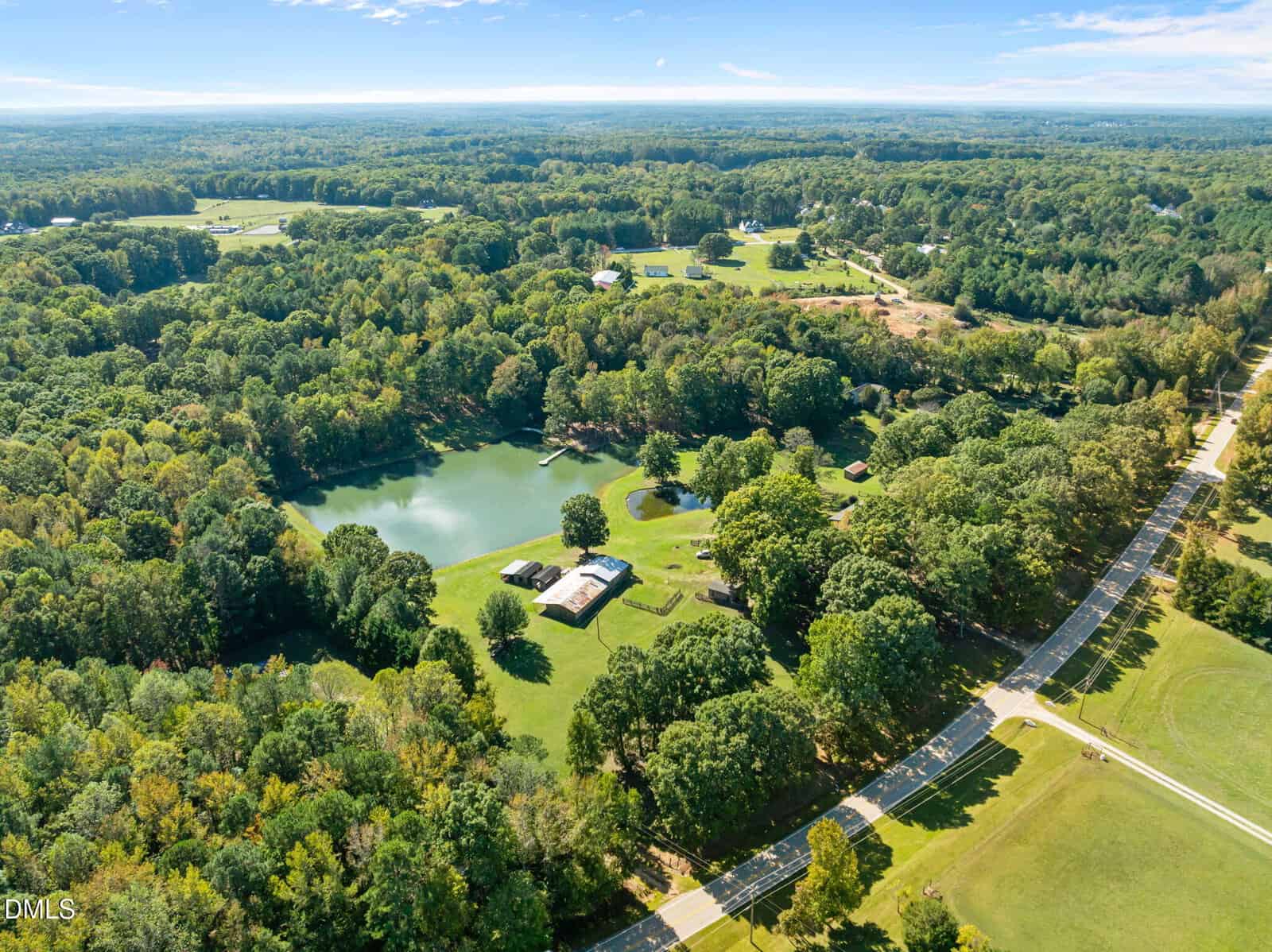 Aerial view of a rural landscape with a pond, several houses, open grassy areas, and tree-lined roads surrounded by dense green forests under a clear blue sky.