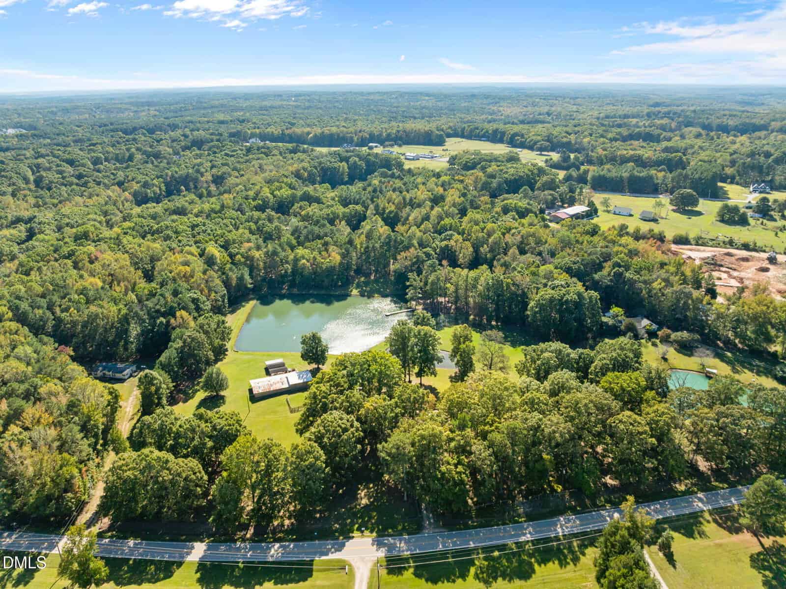 Aerial view of a large, tree-filled property with a pond, a few buildings, grassy open areas, and a road in the foreground, surrounded by dense forests under a clear blue sky.