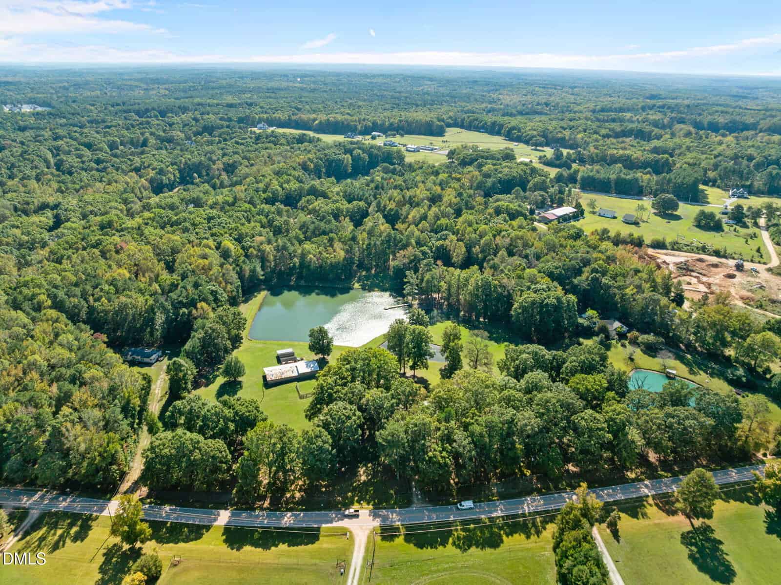 Aerial view of a large, tree-covered area with a central pond, surrounded by open grassy fields, several buildings, and a winding road in the foreground under a clear blue sky.