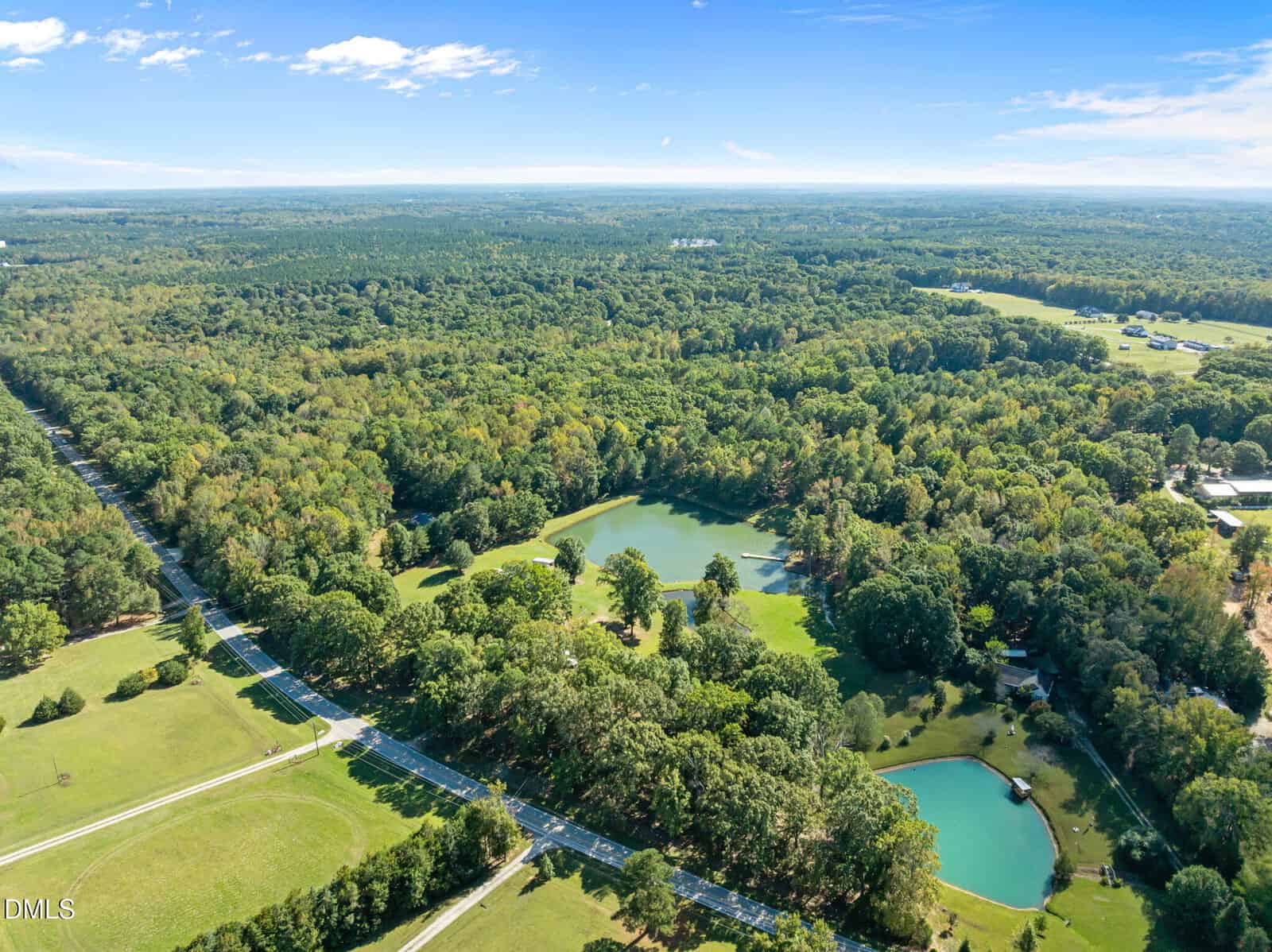 Aerial view of a lush, green landscape with dense forests, two ponds, open grassy areas, and a road running alongside the trees under a blue sky with scattered clouds.