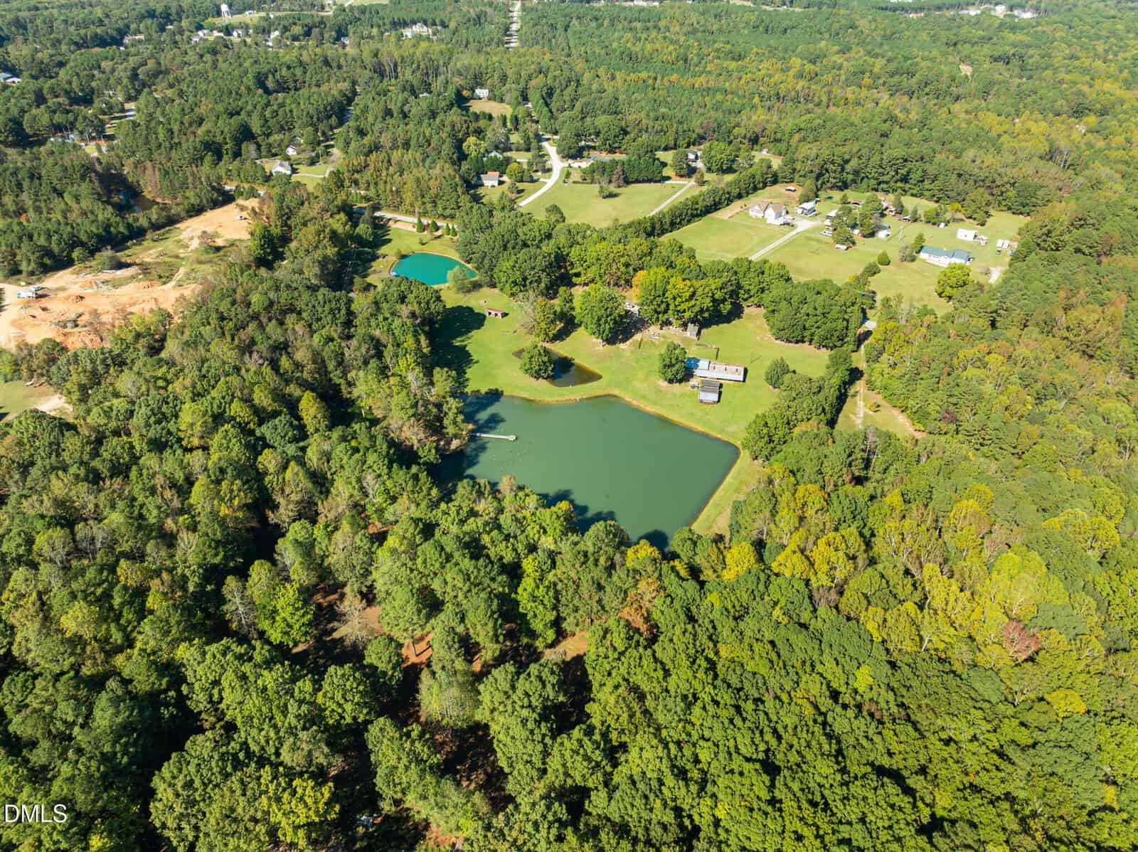 Aerial view of a rural landscape featuring dense green trees, two small ponds, scattered houses, open grassy lawns, and winding roads under a clear sky.